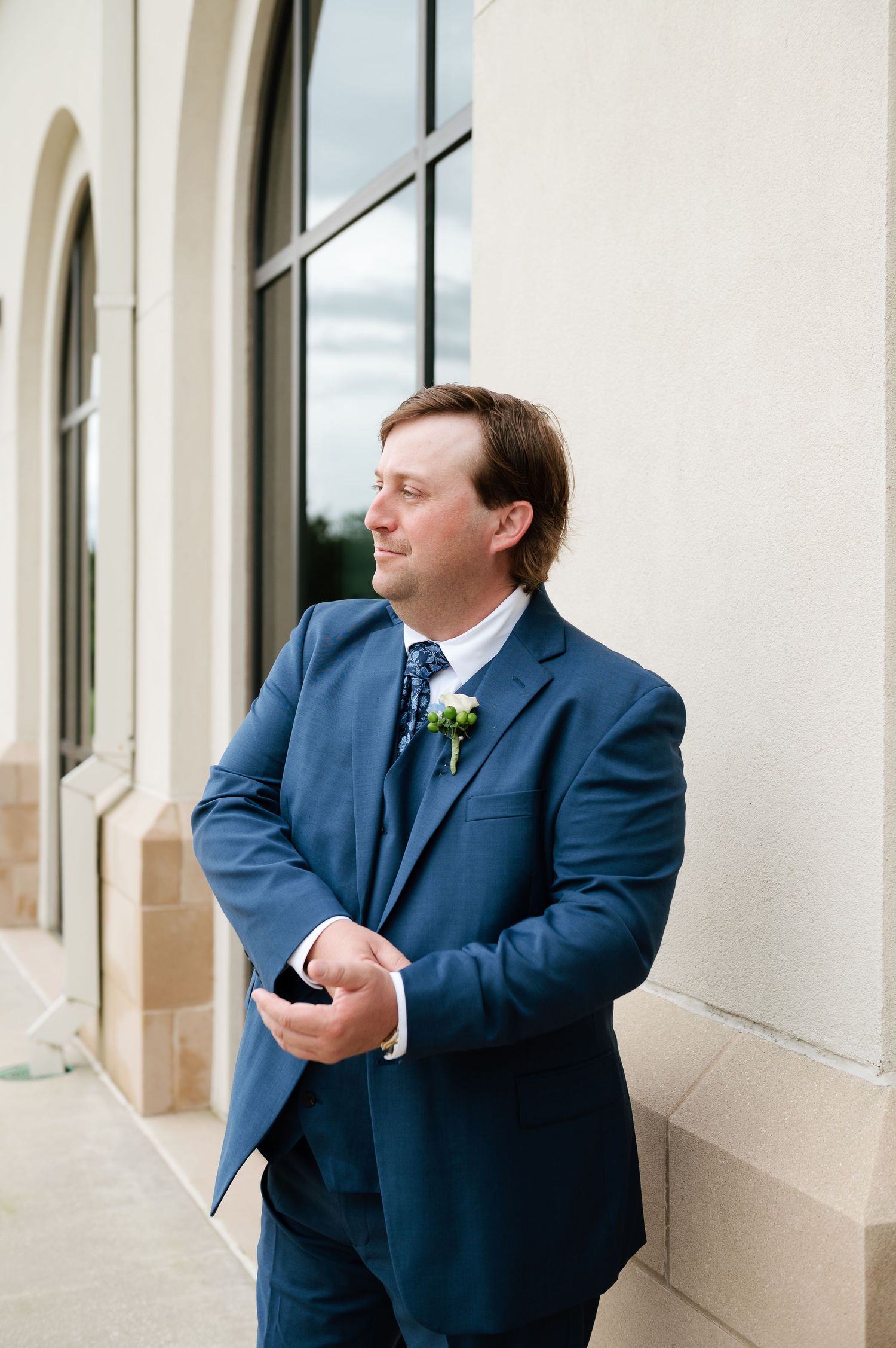 Groom in navy blue suit with boutonniere holds phone while standing against stone building column.