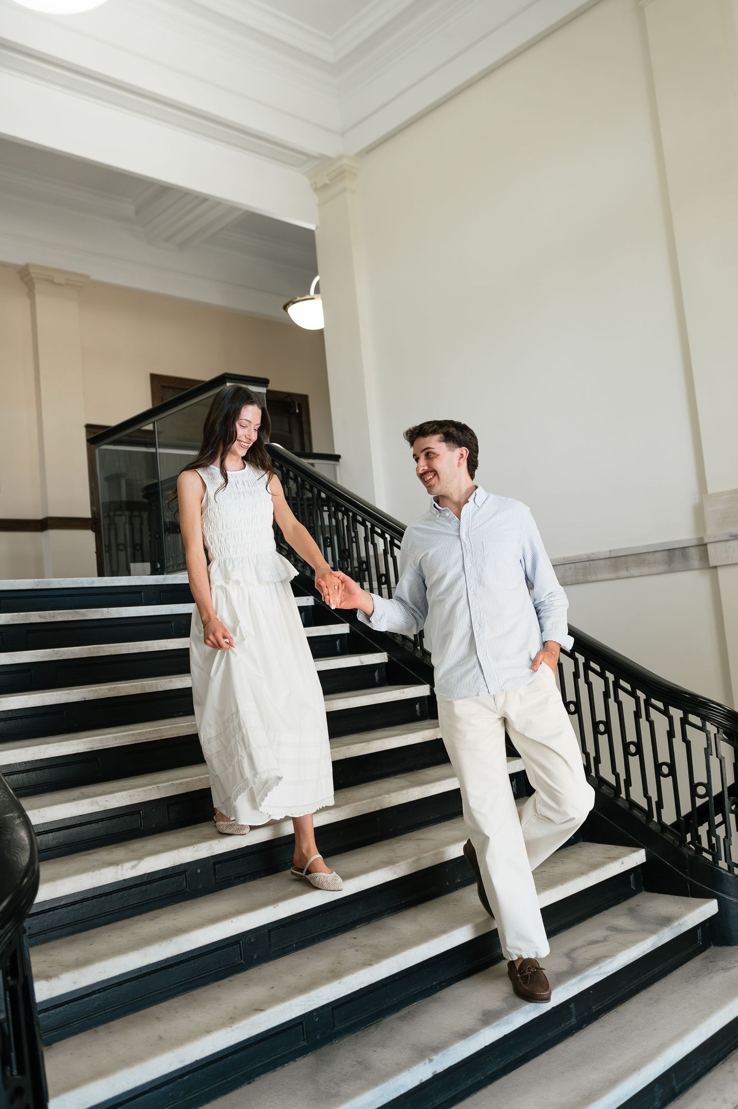 A couple holds hands while walking down elegant marble stairs with black railings in a bright classical building.