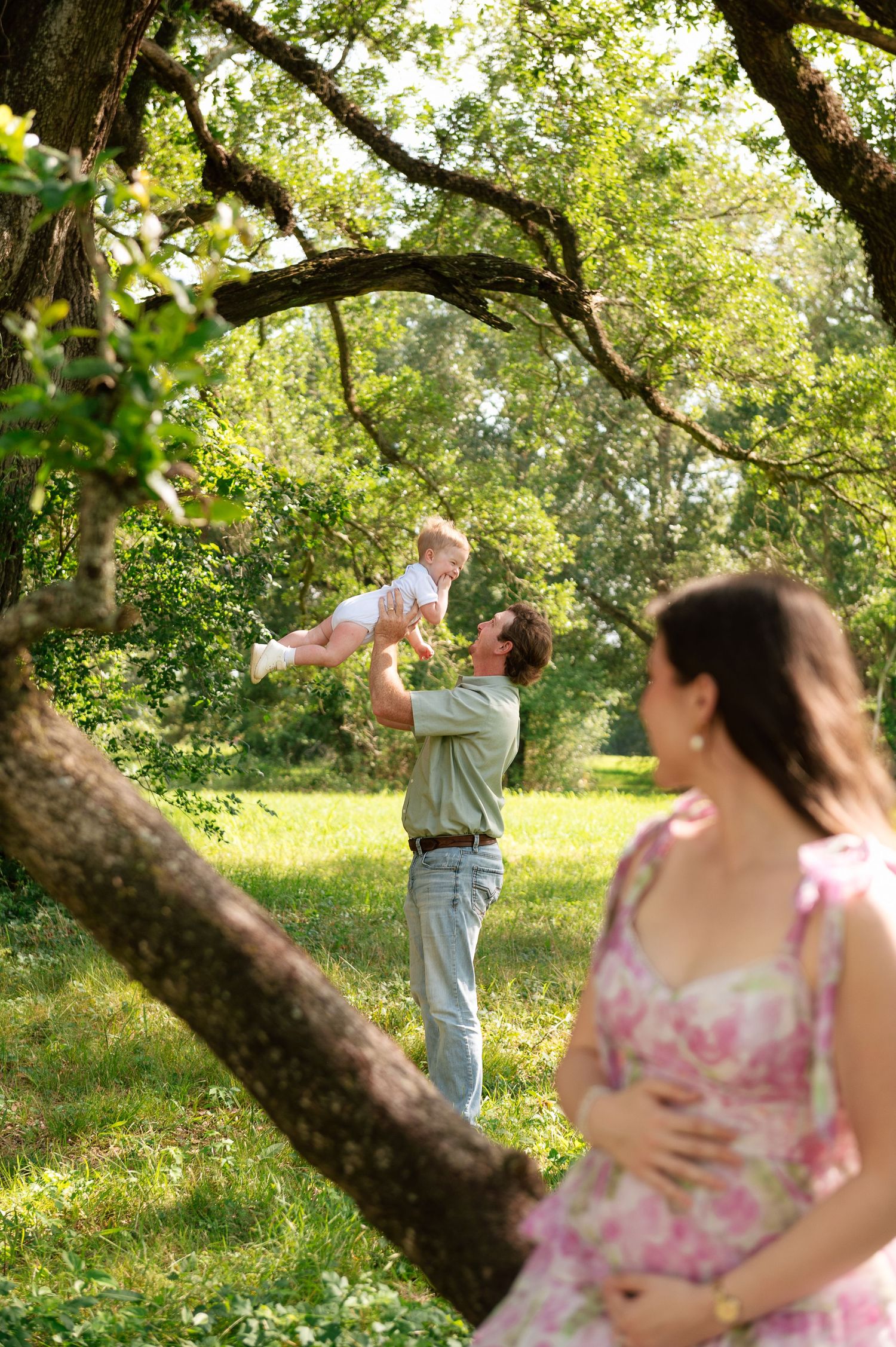 Pregnant woman in floral dress watches man lift baby in sunny green park.