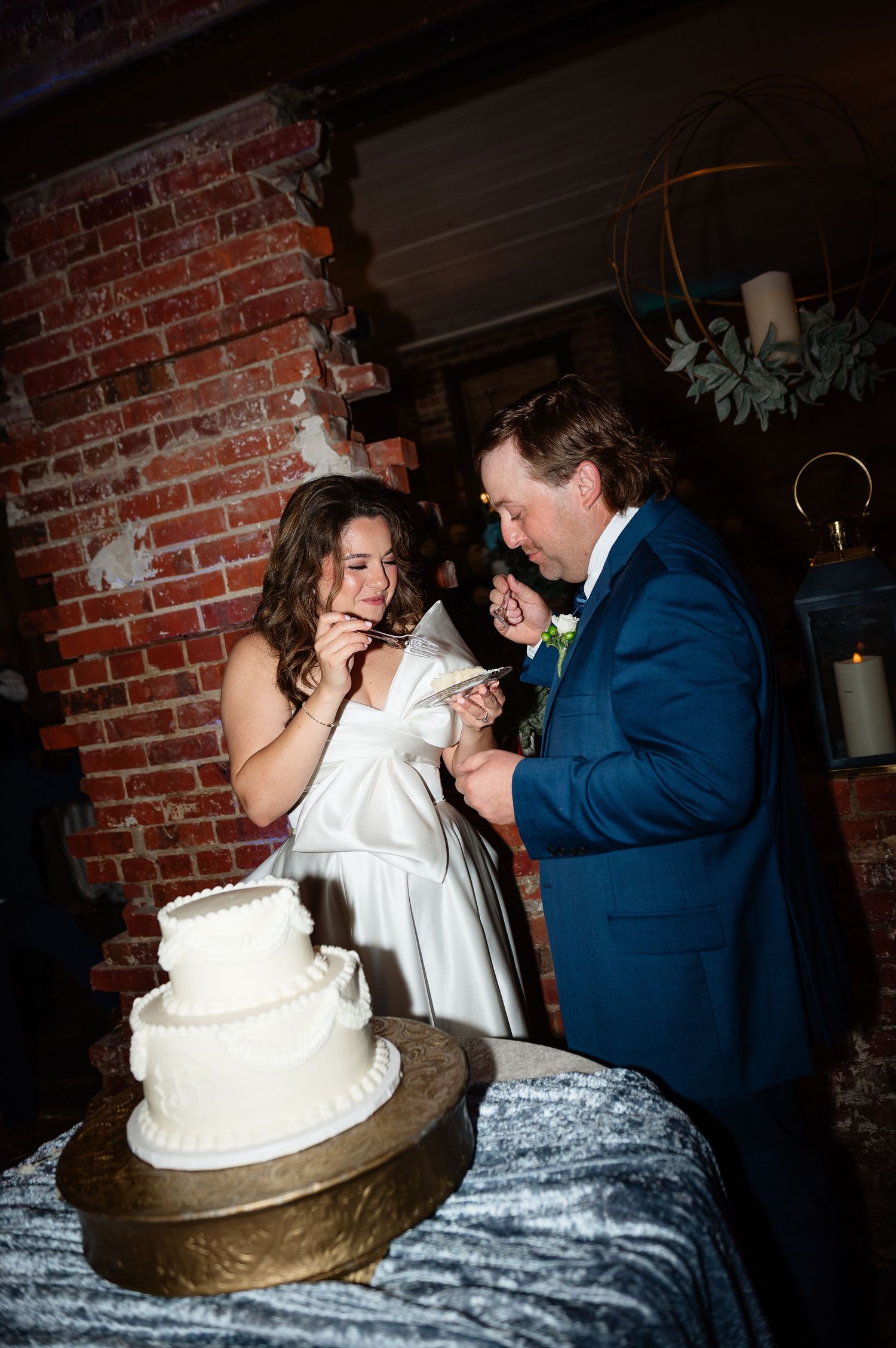 Bride and groom laughing while feeding each other wedding cake at rustic brick venue reception.