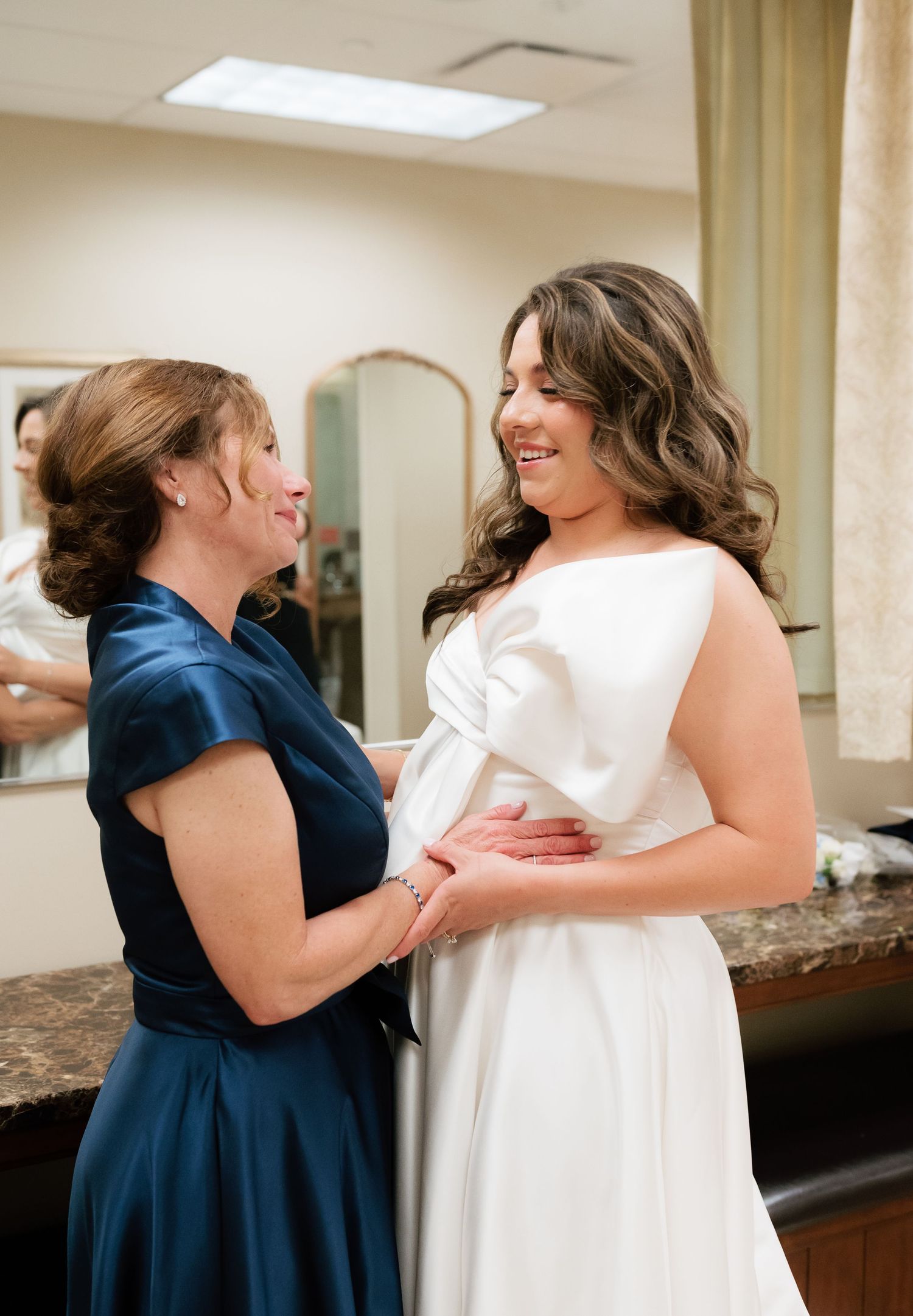 Bride in white gown smiling with woman in navy blue dress during wedding preparations in bridal suite.