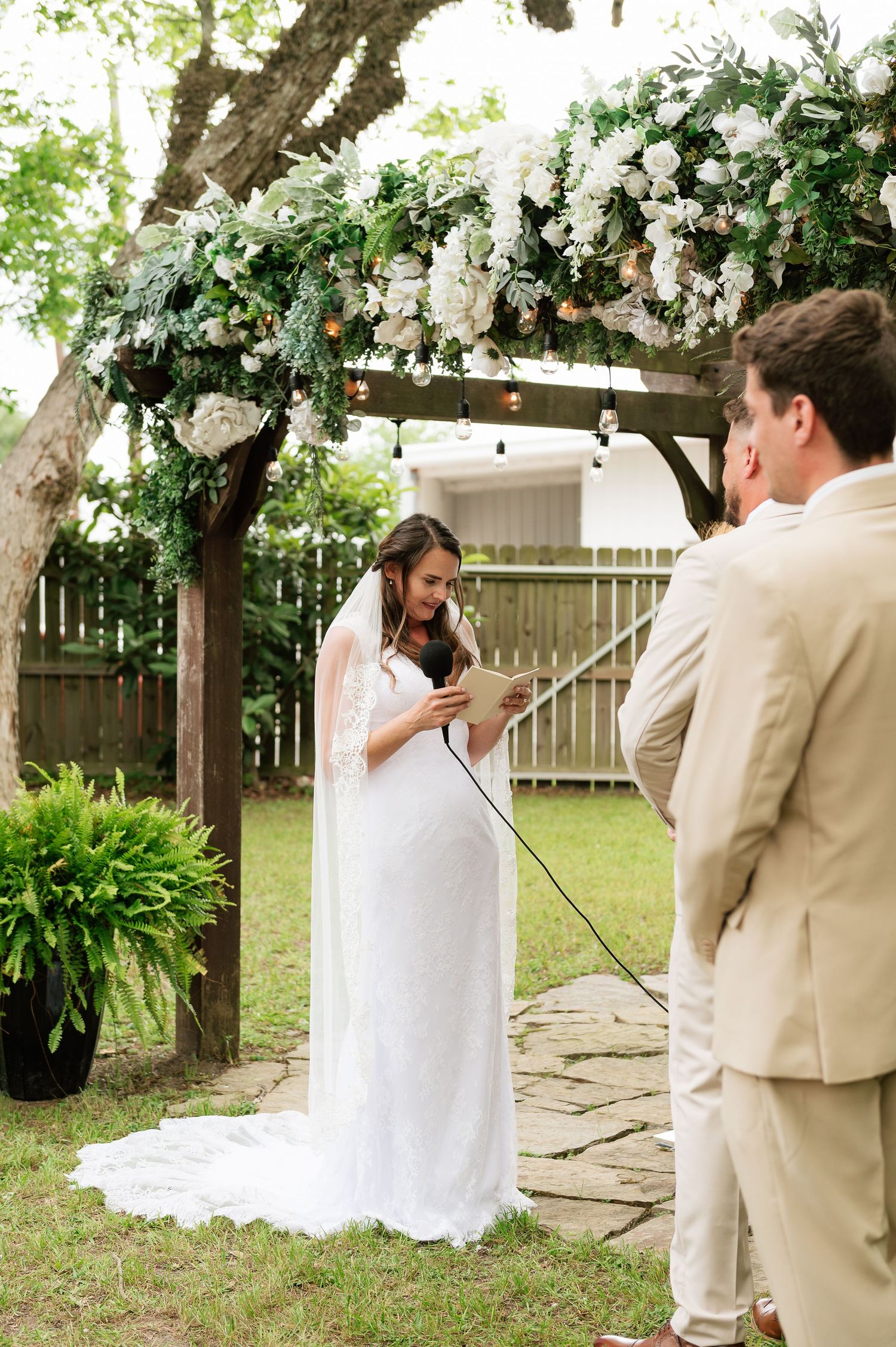 Bride in white gown reads vows under floral arch with greenery at outdoor garden wedding ceremony.