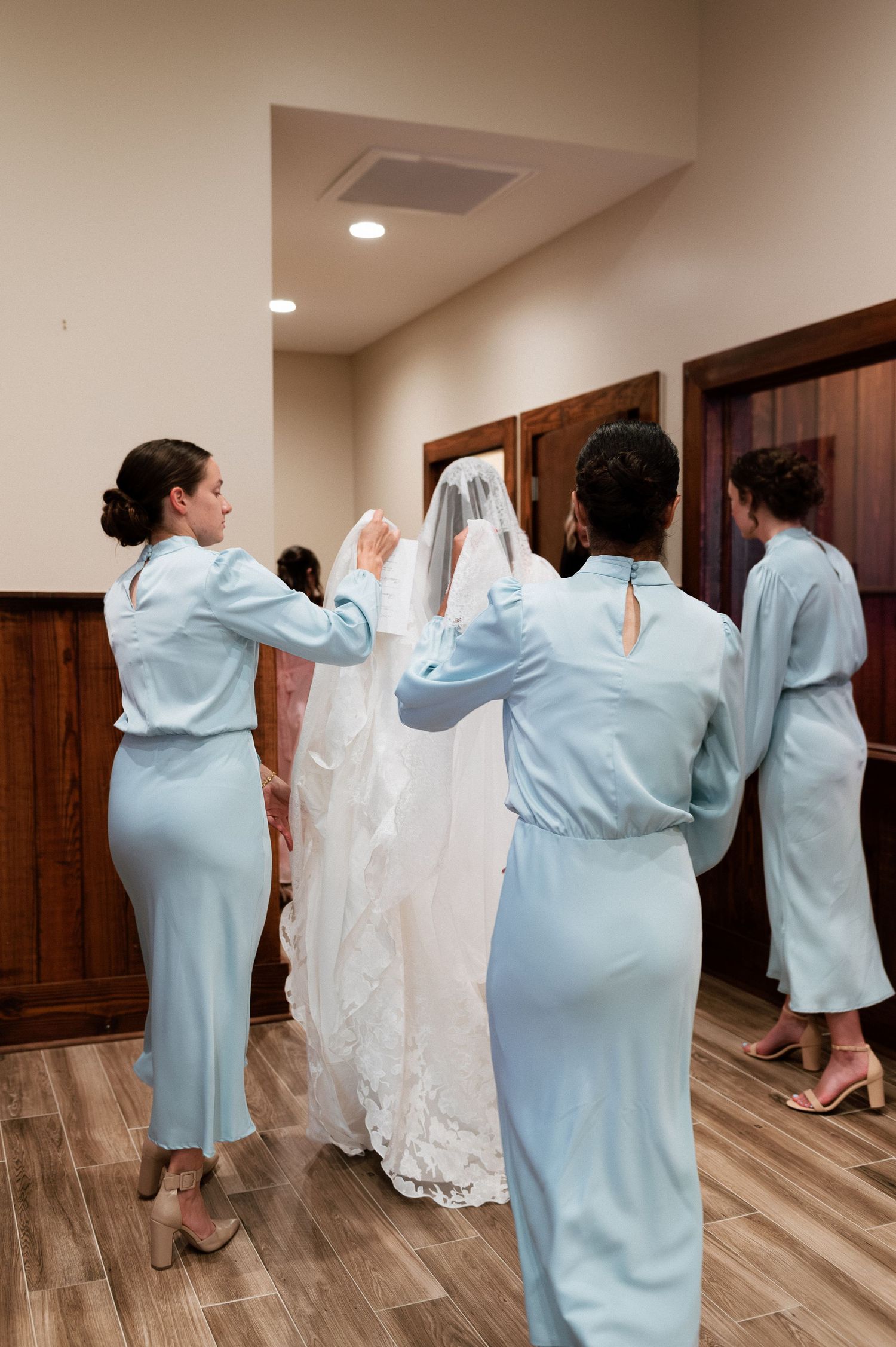 Bridesmaids in light blue dresses help bride with wedding veil in elegant venue hallway.