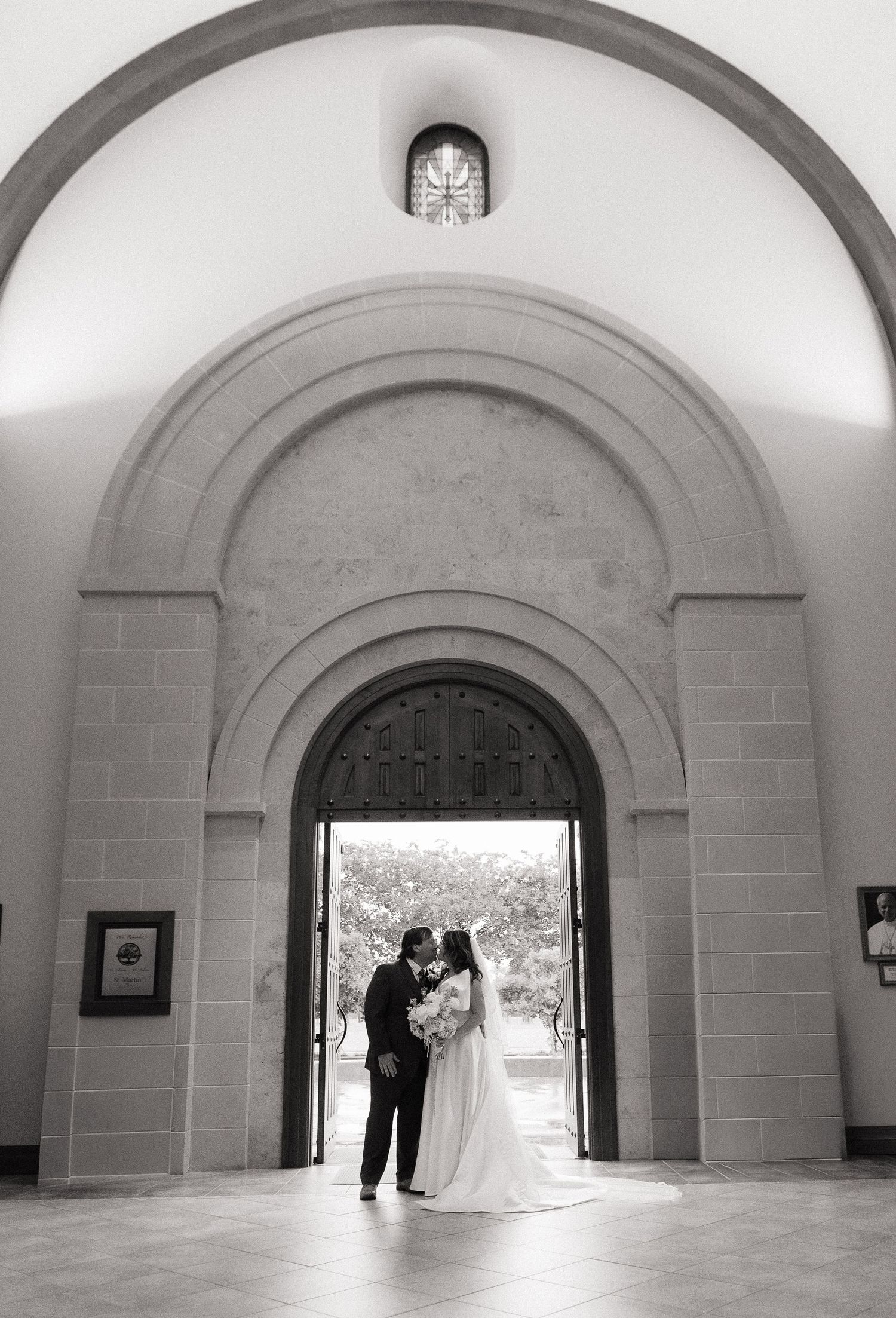 A bride and groom share a romantic moment under an elegant stone arch doorway in a black and white wedding photo.
