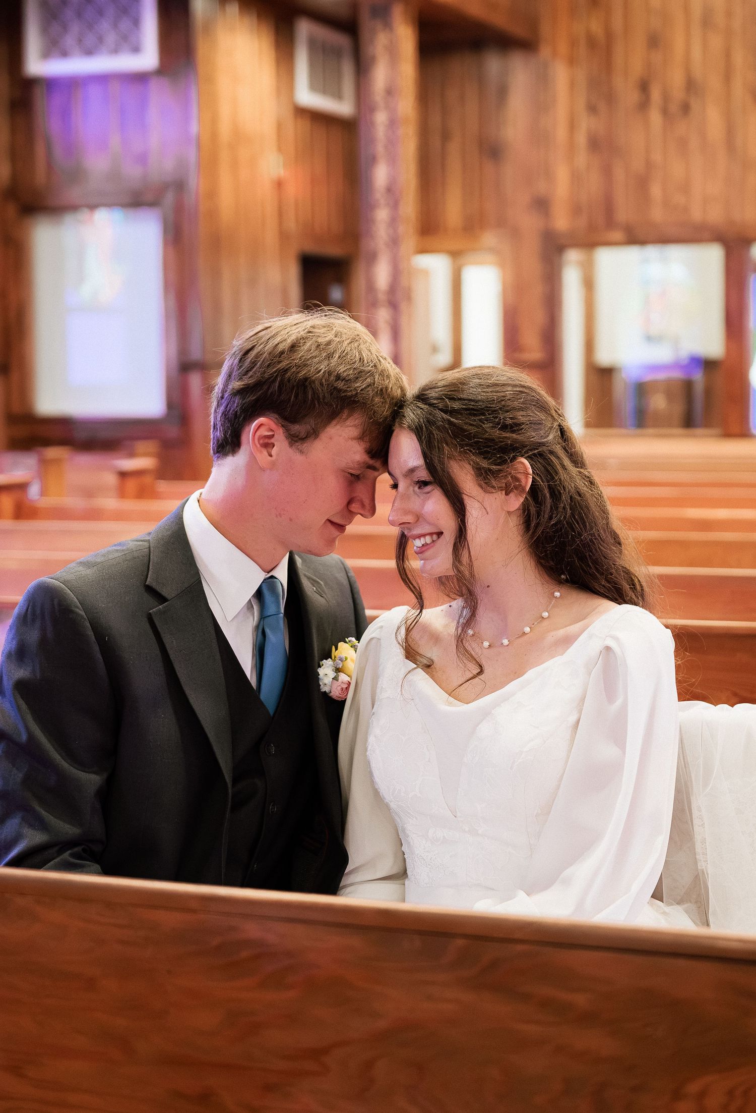 Bride and groom share a romantic forehead touch while seated in a wooden church pew on their wedding day.