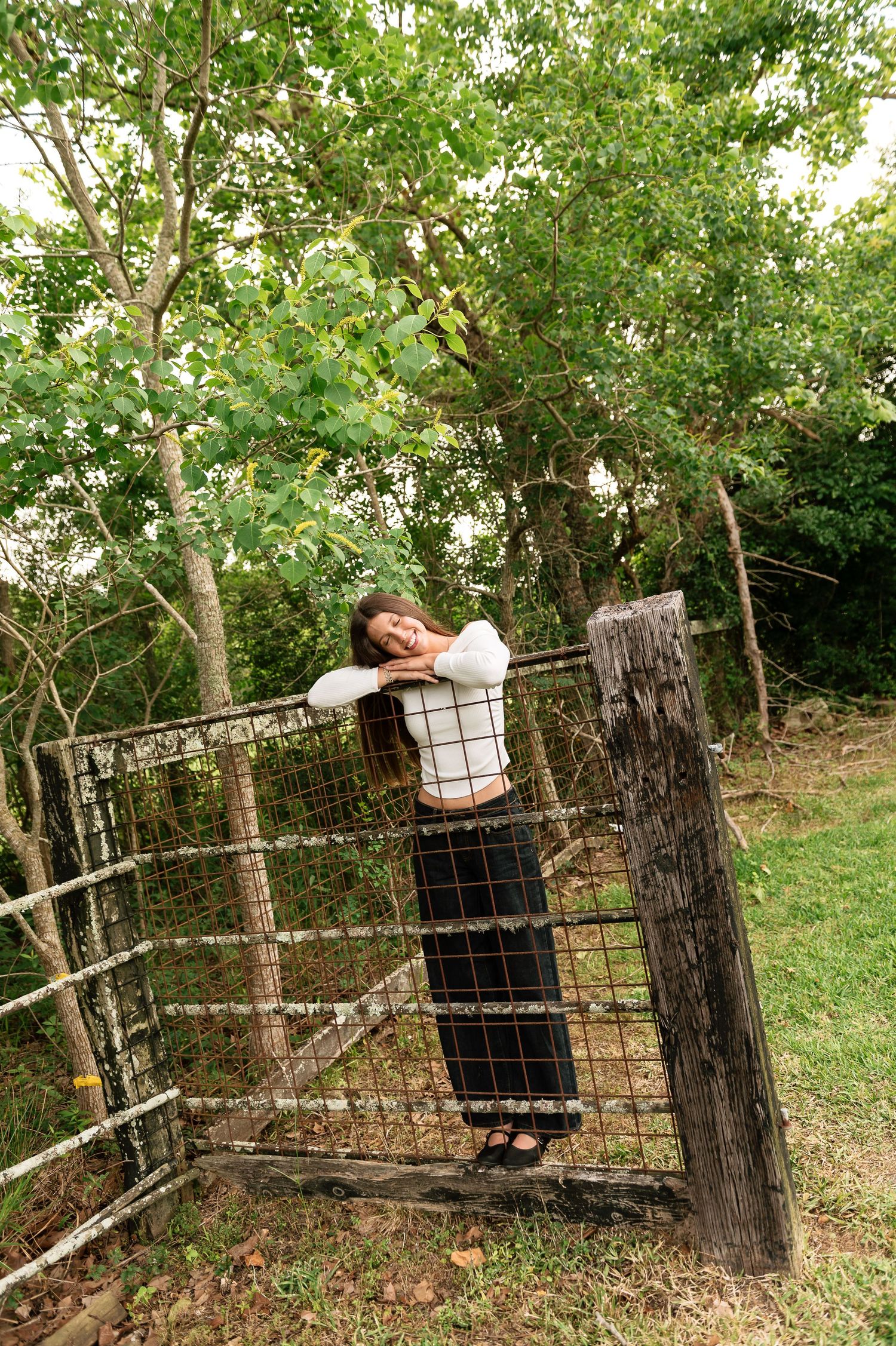 Woman in white top and black skirt leaning on rustic metal farm gate surrounded by lush green trees.