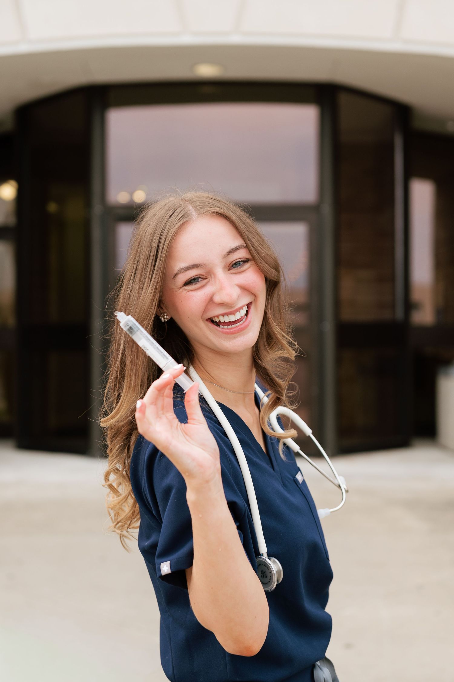 Smiling nursing student in navy scrubs holding a syringe with stethoscope outside a medical building.
