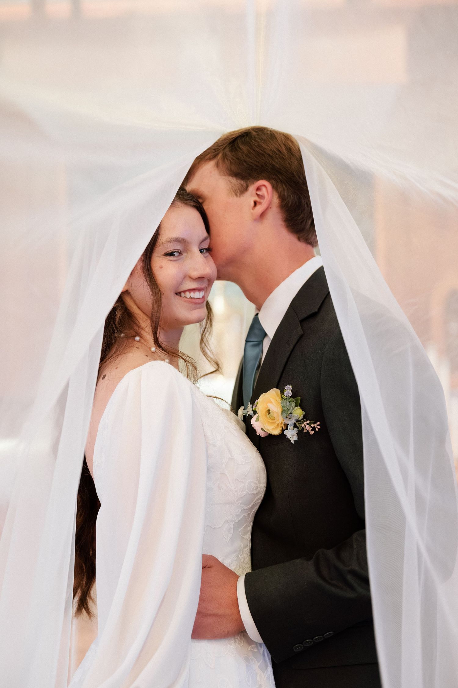 Bride and groom share a romantic moment under a flowing white veil, with the groom kissing her cheek at their wedding.