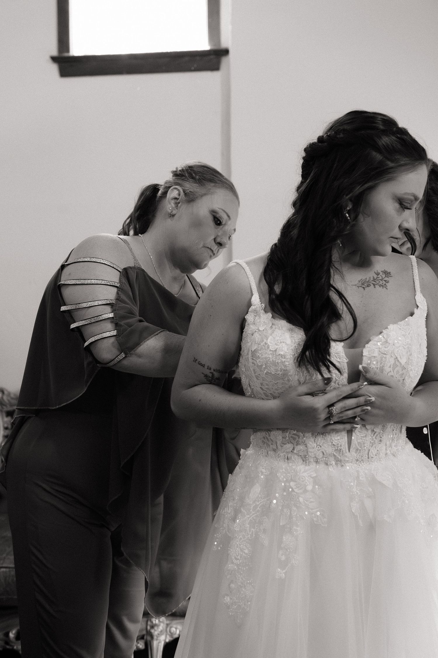 Black and white photo of a bride in lace gown being helped by bridesmaid in elegant dress during wedding preparations.