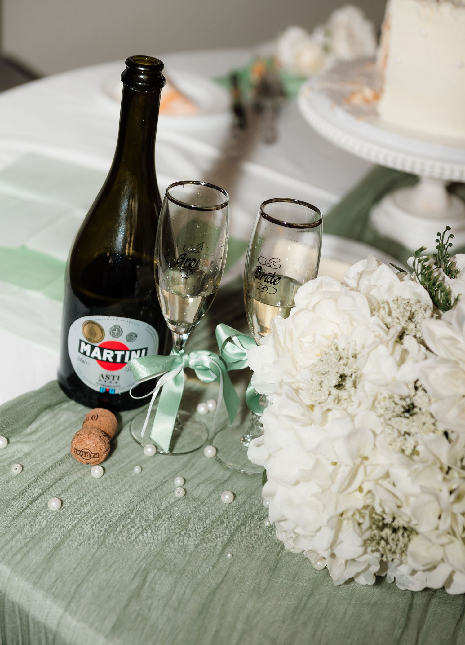 Martini Asti bottle with champagne flutes and white floral bouquet on mint green table runner at wedding reception.