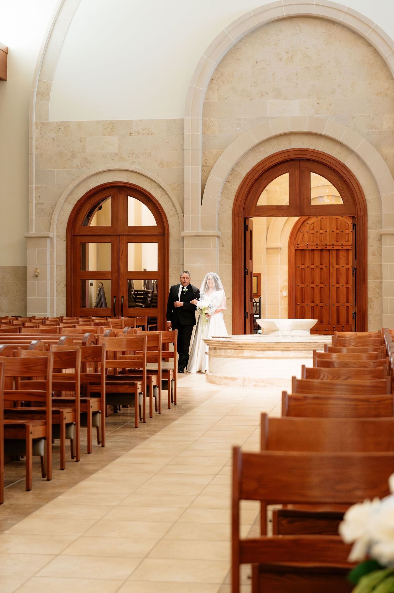 Bride and escort walk down aisle of elegant stone church with wooden pews and arched doorways.