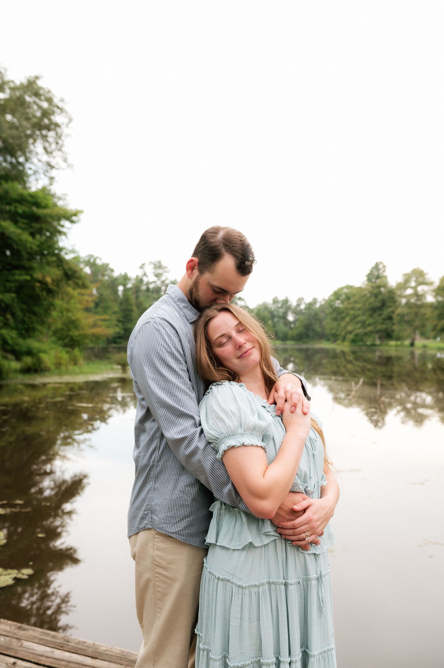 Couple embracing tenderly on a wooden dock by a serene lake, woman in mint dress, man in gray shirt, surrounded by lush trees.