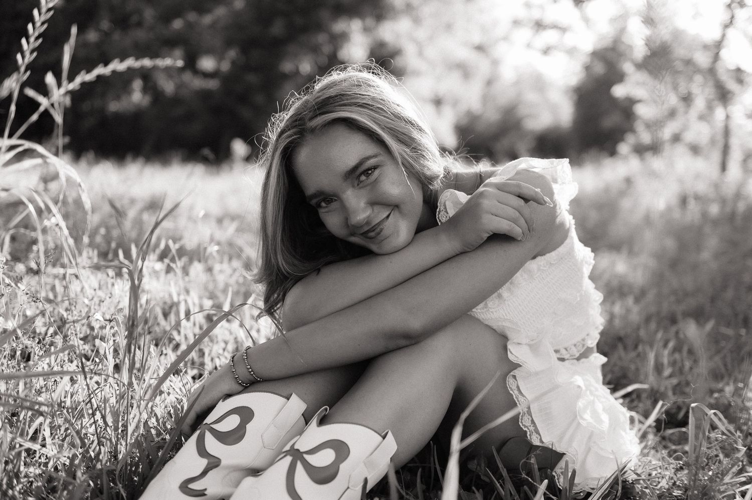 Smiling young woman in a white dress and cowboy boots sits hugging her knees in a sunlit grassy field in a black and white portrait.