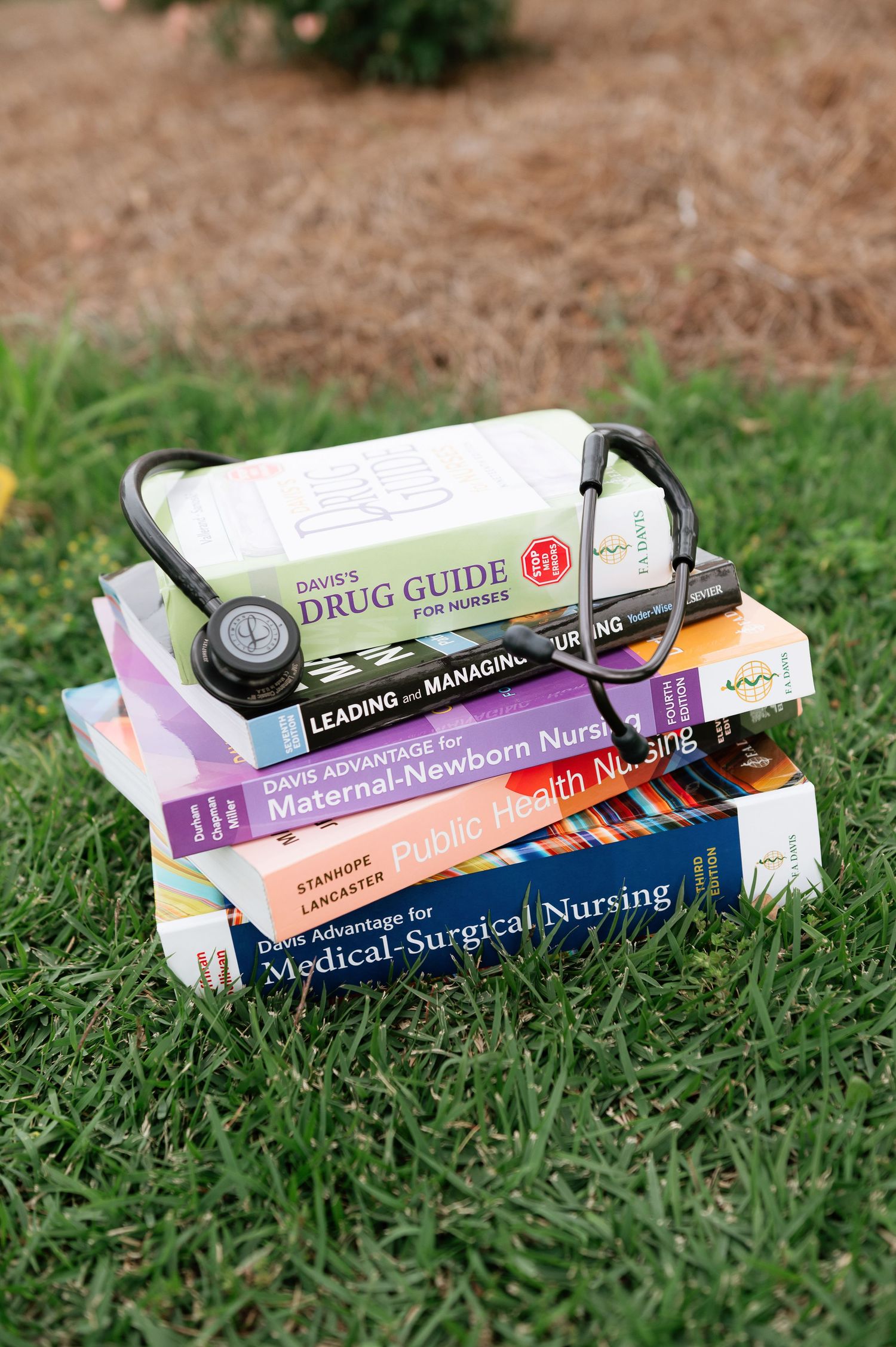 Stack of nursing textbooks with stethoscope on grass, including Drug Guide, Public Health and Medical-Surgical Nursing books.
