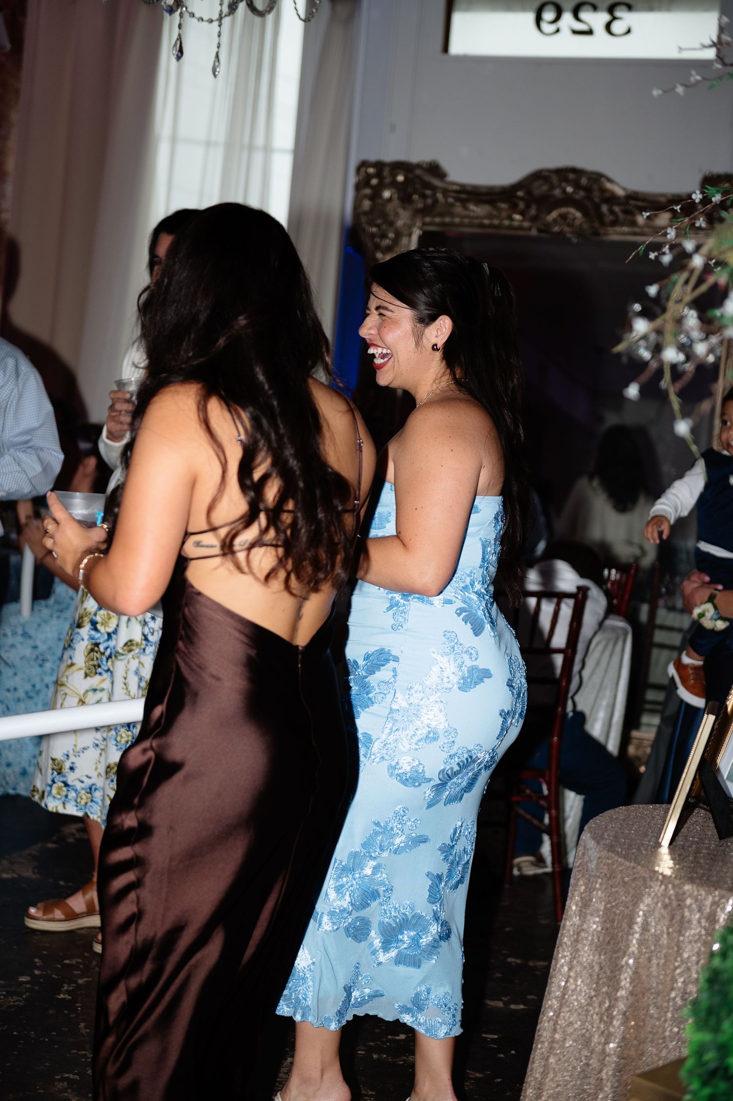 Two women in elegant dresses laughing and dancing together at a wedding reception celebration.
