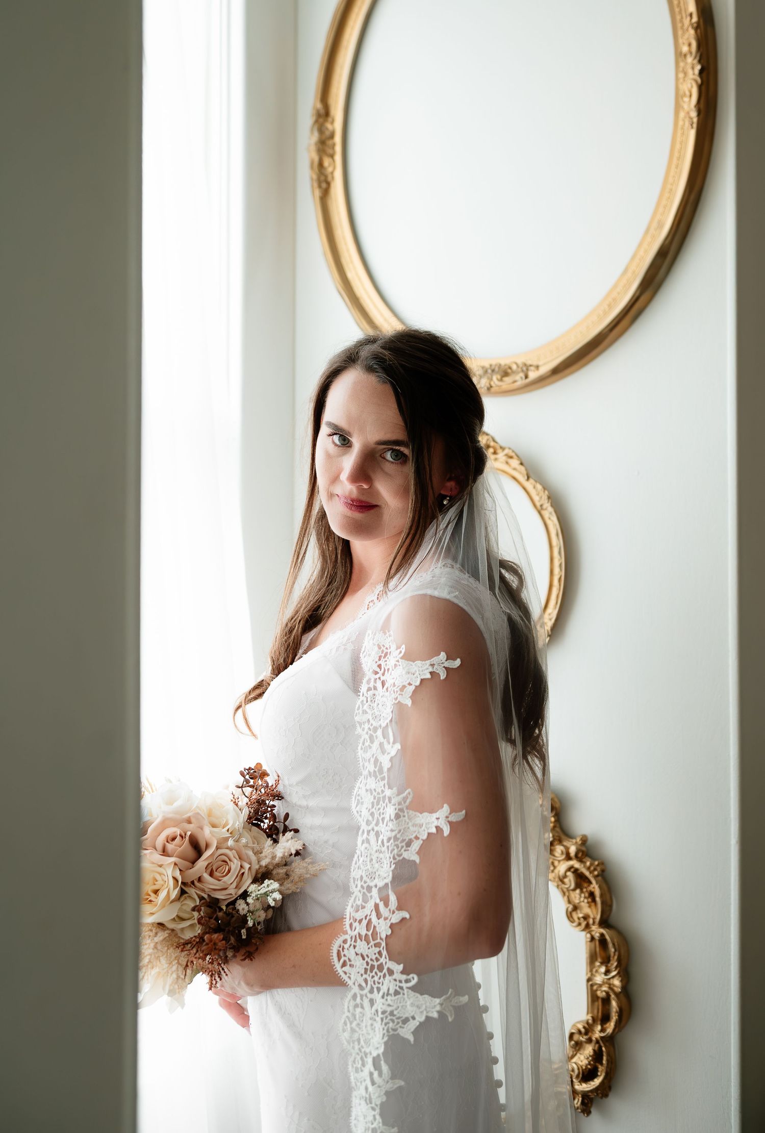 Bride in white lace gown and veil holds bouquet by golden mirrors.