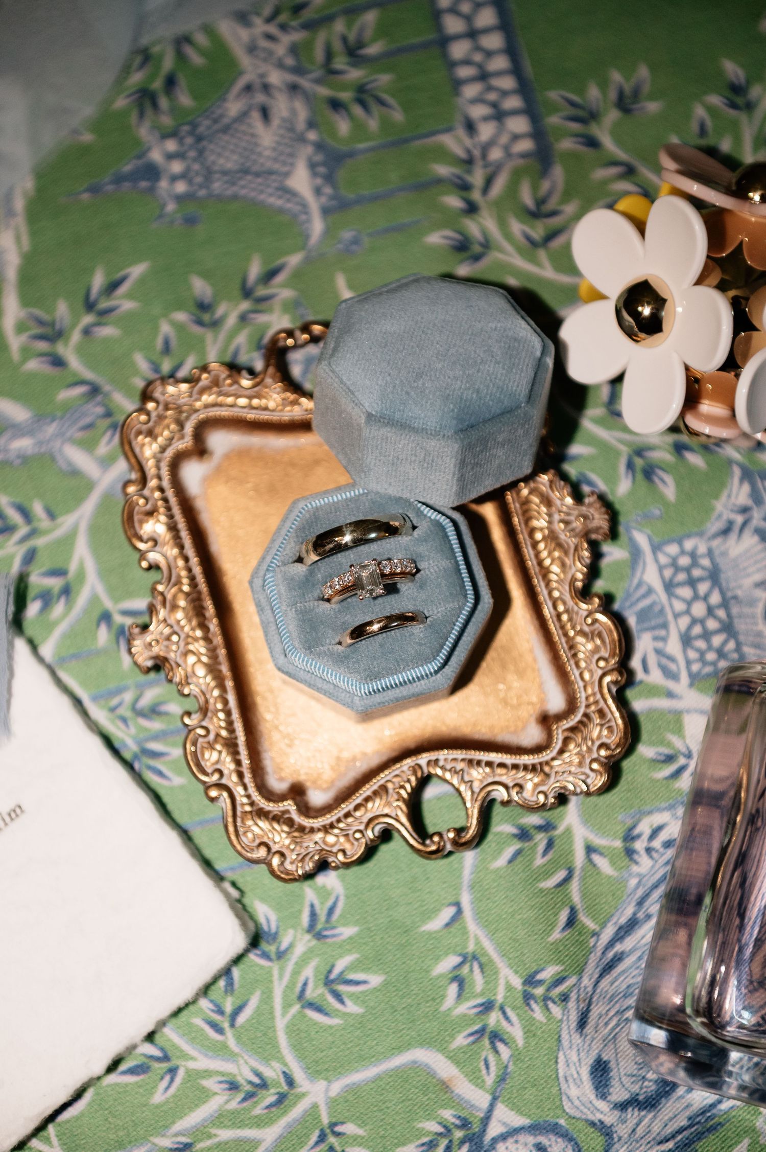 Blue velvet ring box with wedding bands on gold ornate tray atop green floral tablecloth.