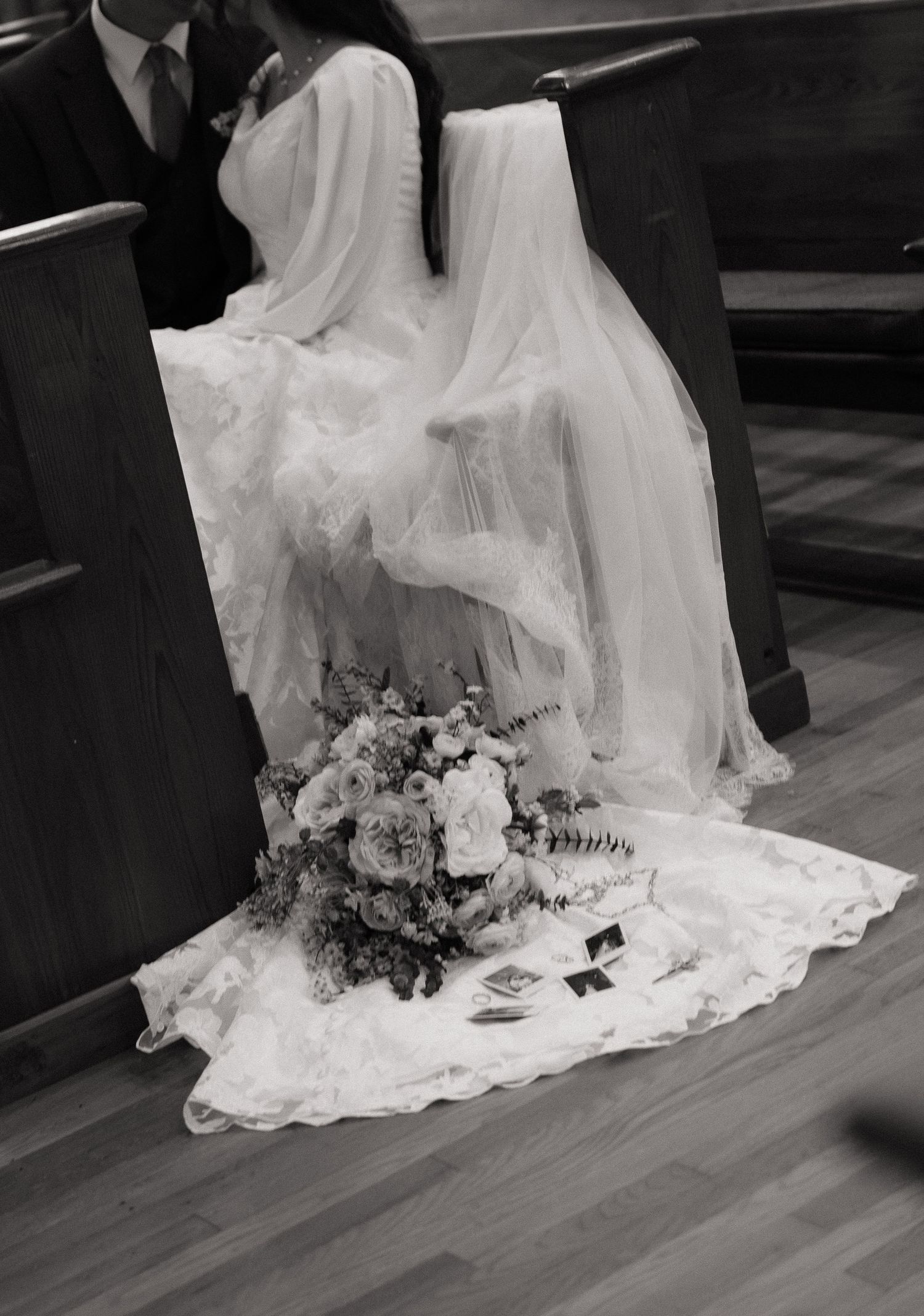 Black and white wedding photo of bride kneeling at church pew with bouquet and veil train spread on floor.
