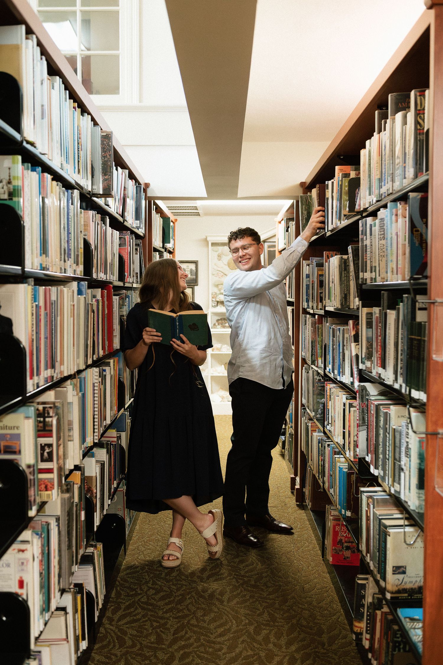 A couple browses books together in a library aisle, woman holding open book, man reaching for shelf.