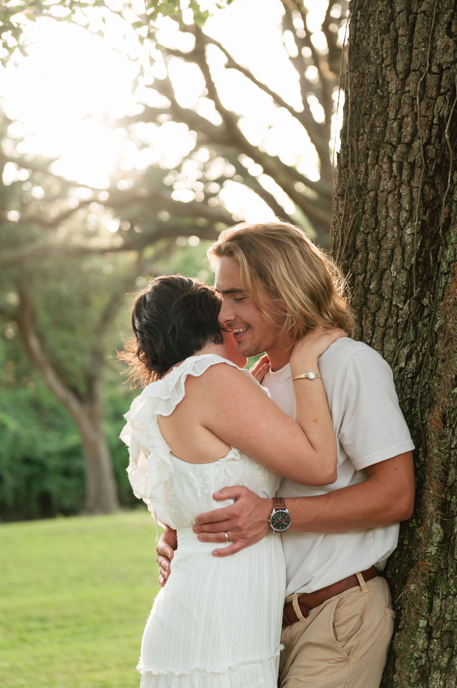 A smiling couple embraces romantically by a tree in a sunlit park, woman in white dress, man in casual attire.