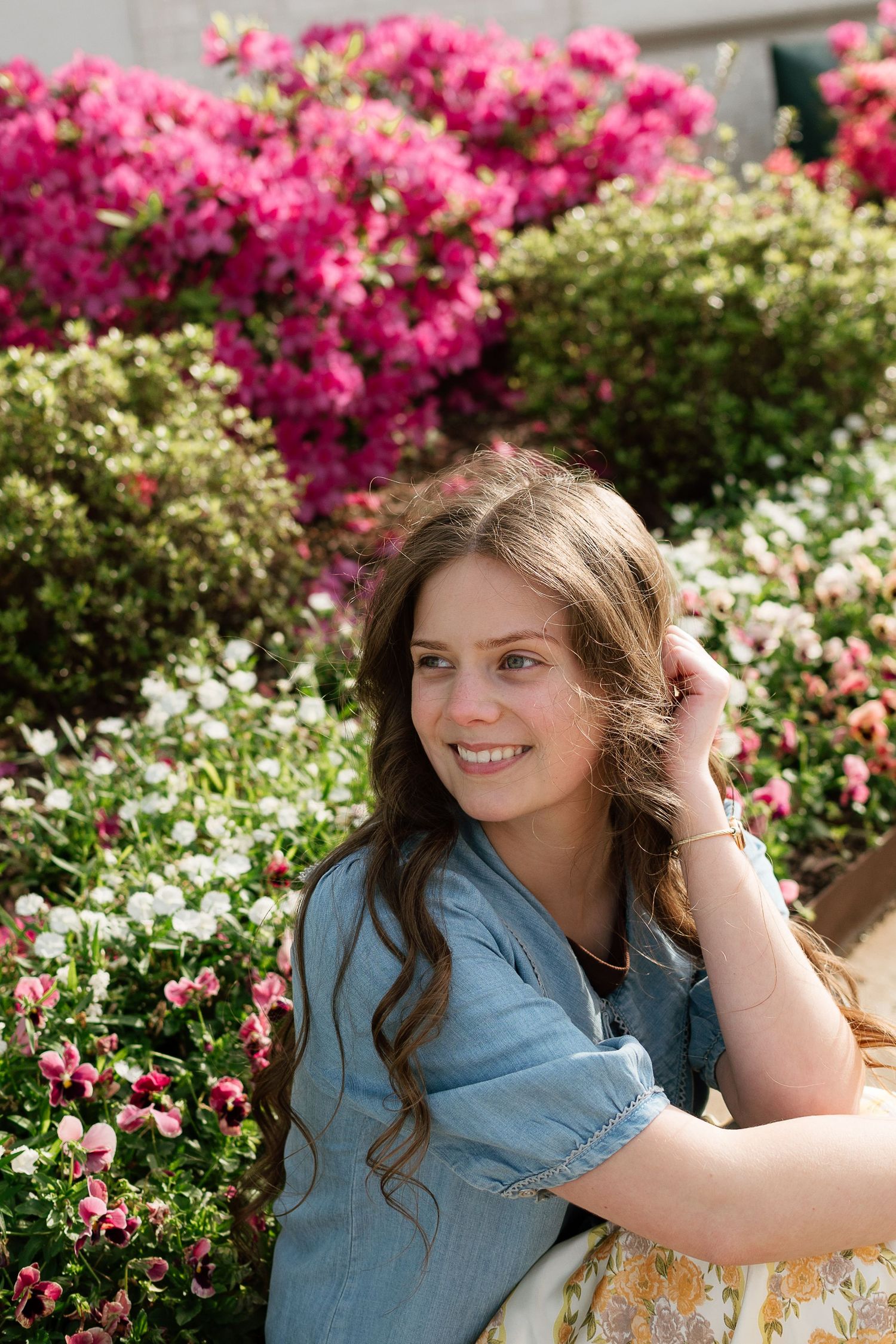 Smiling young woman in denim shirt poses among vibrant pink and white flowers in a sunny garden.