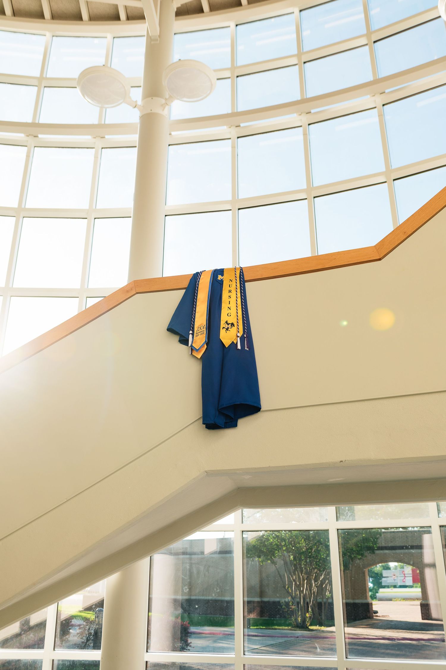 Blue graduation gown and gold honor stole draped over a staircase railing in a bright, modern building atrium.