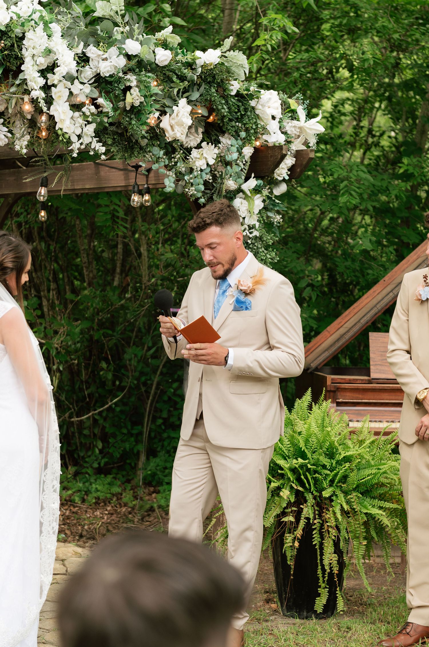 Groom in tan suit reads vows from brown book at outdoor wedding ceremony beneath floral arch.