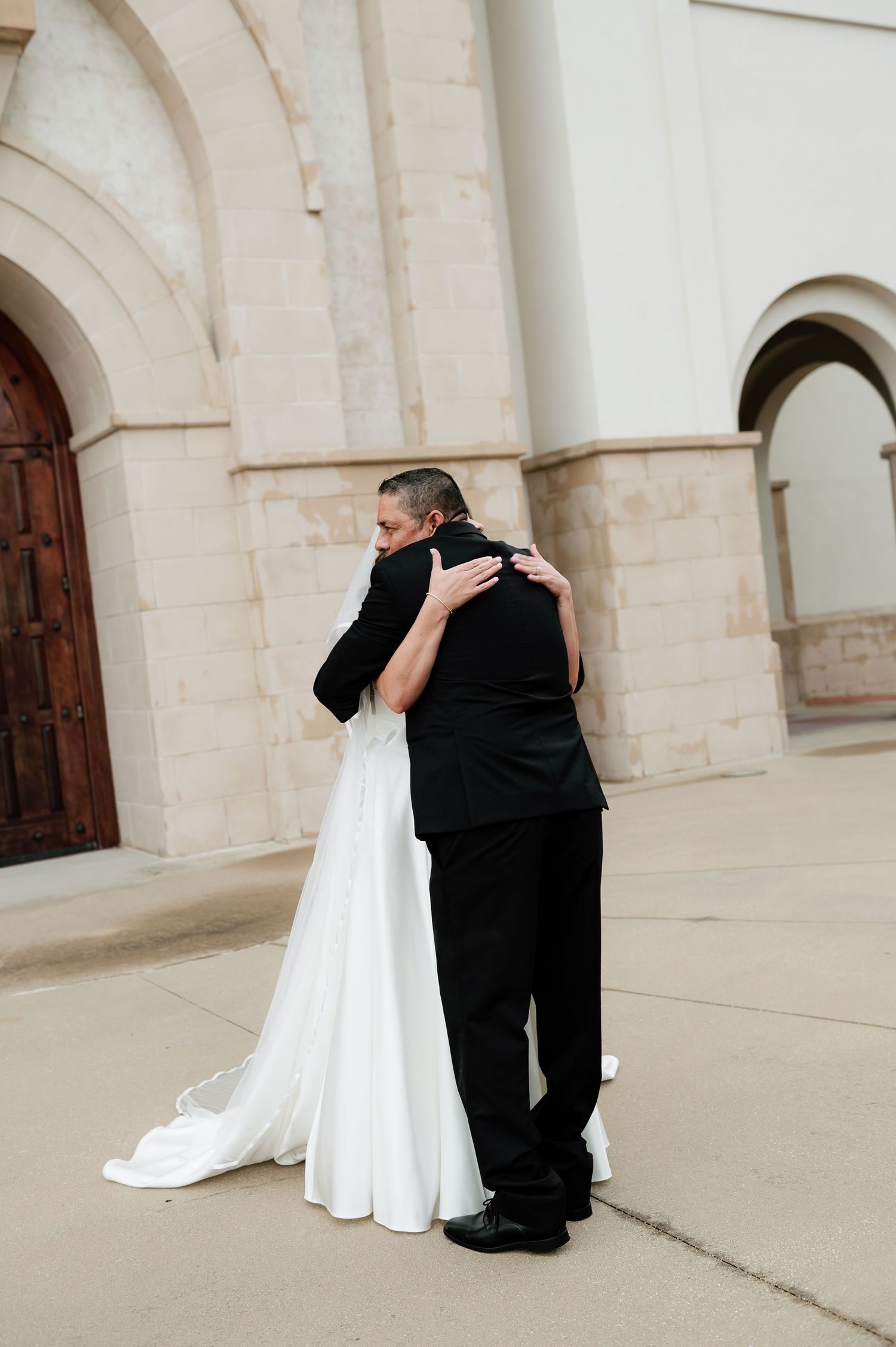 Bride and groom sharing emotional first look hug outside stone church with arched wooden doors.