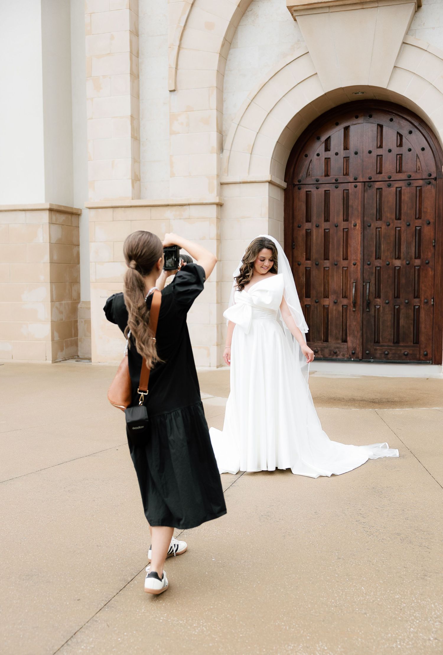 Photographer in black dress captures bride in white gown with veil posing before large wooden doors of stone church.