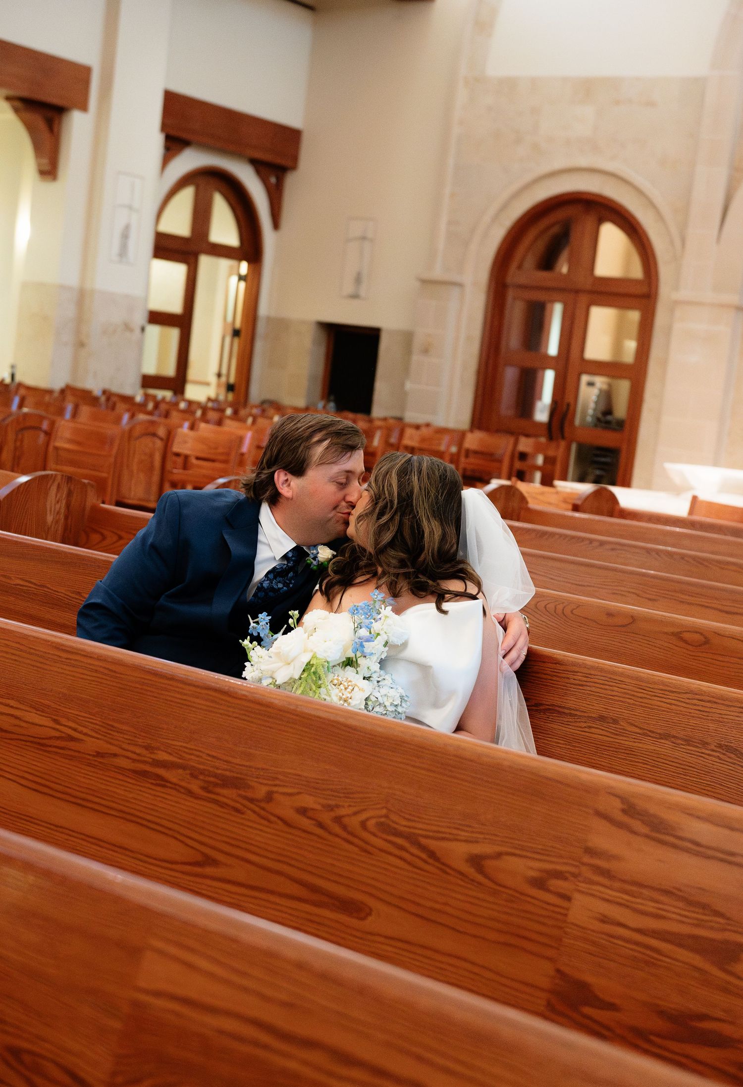 Bride and groom sharing a kiss among wooden church pews during their wedding ceremony.