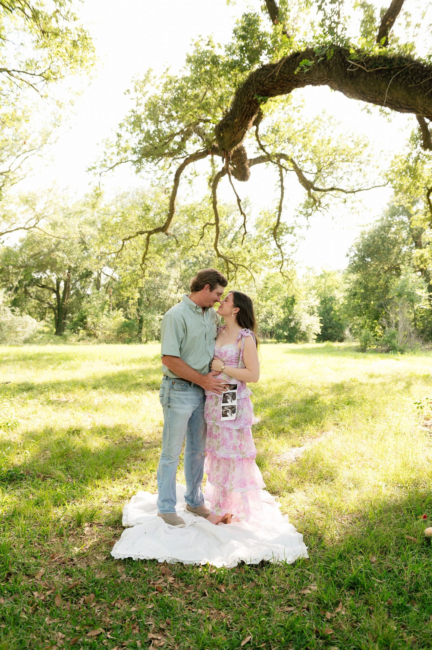 Couple sharing a tender moment under a sprawling oak tree in a lush green park, holding an ultrasound photo.