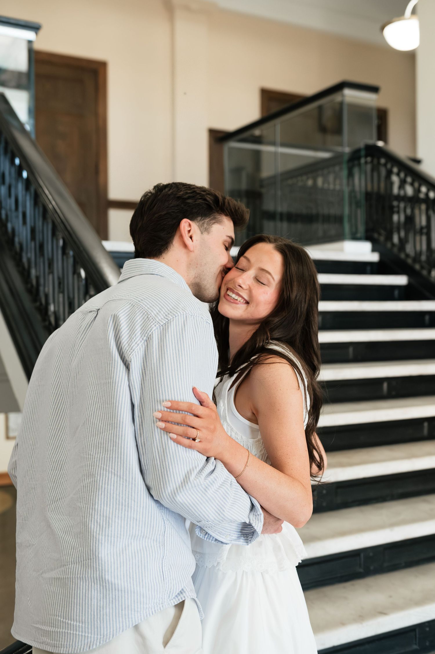 Couple embracing and smiling near a staircase, man kissing woman on cheek, woman wearing white dress.
