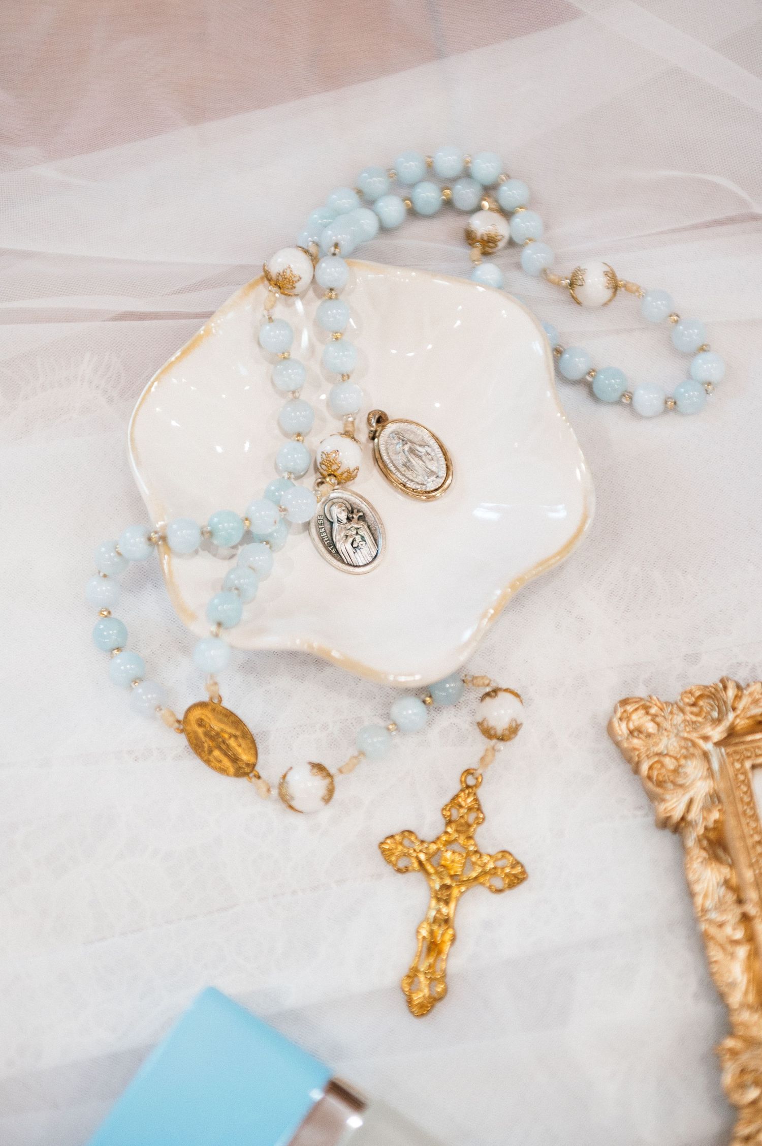 Light blue rosary beads, gold cross, religious medals, and rings arranged on a white decorative dish.