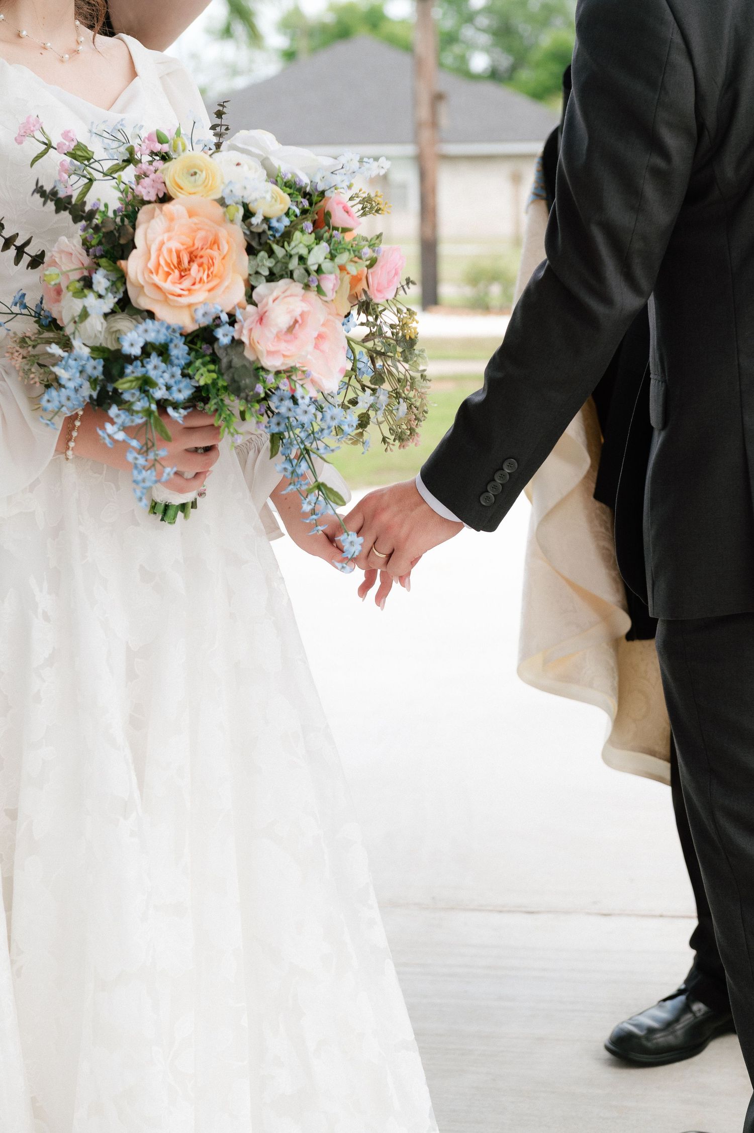 Bride and groom holding hands, bride wearing white lace gown holding pastel bouquet of peach roses and blue flowers.
