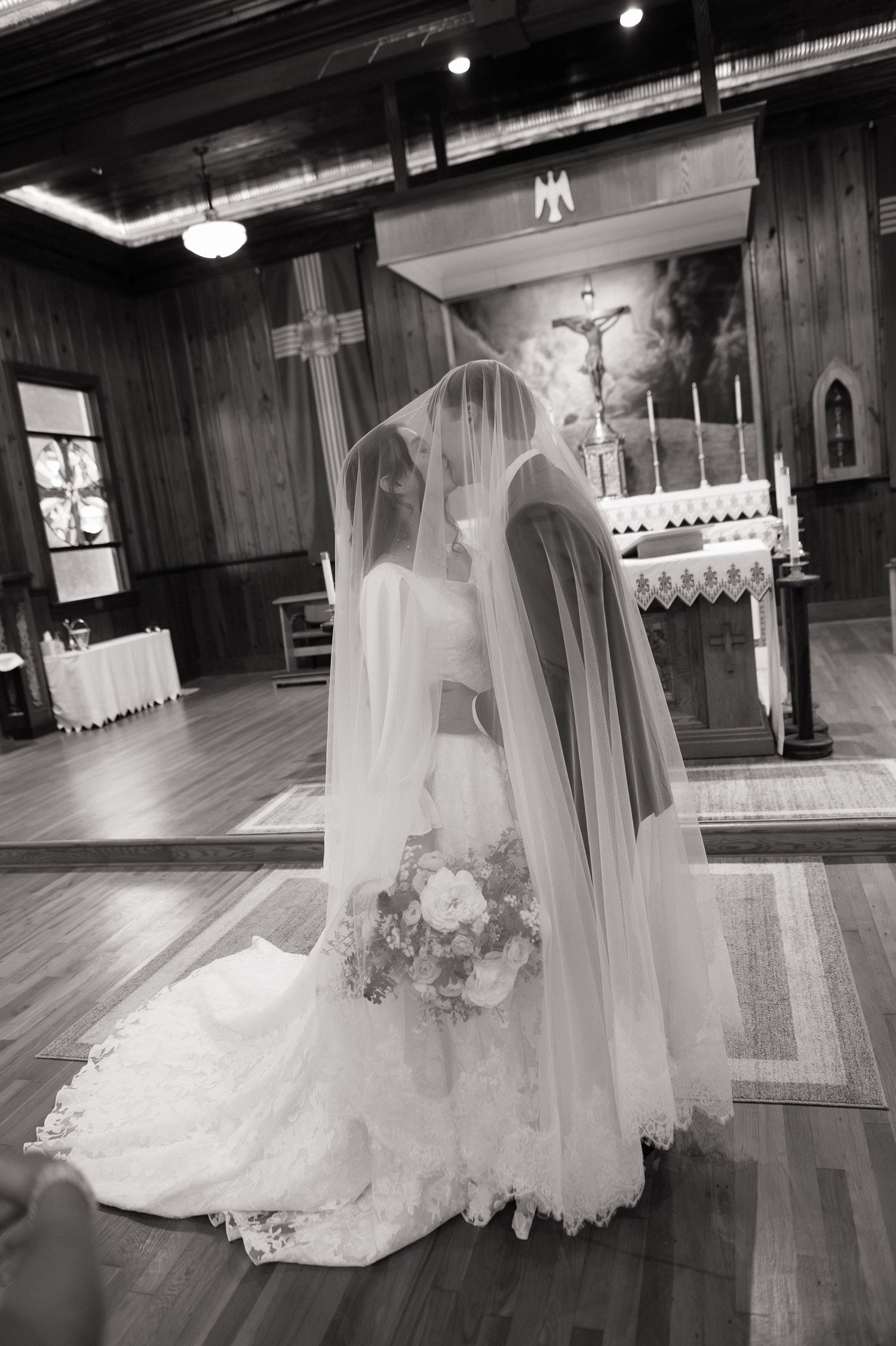 Black and white photo of a bride in a flowing veil and lace gown holding a bouquet at a church altar.