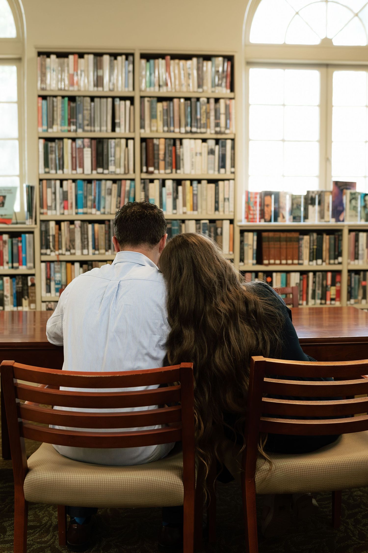 Couple sitting together at a library table, leaning close, surrounded by bookshelves filled with books.