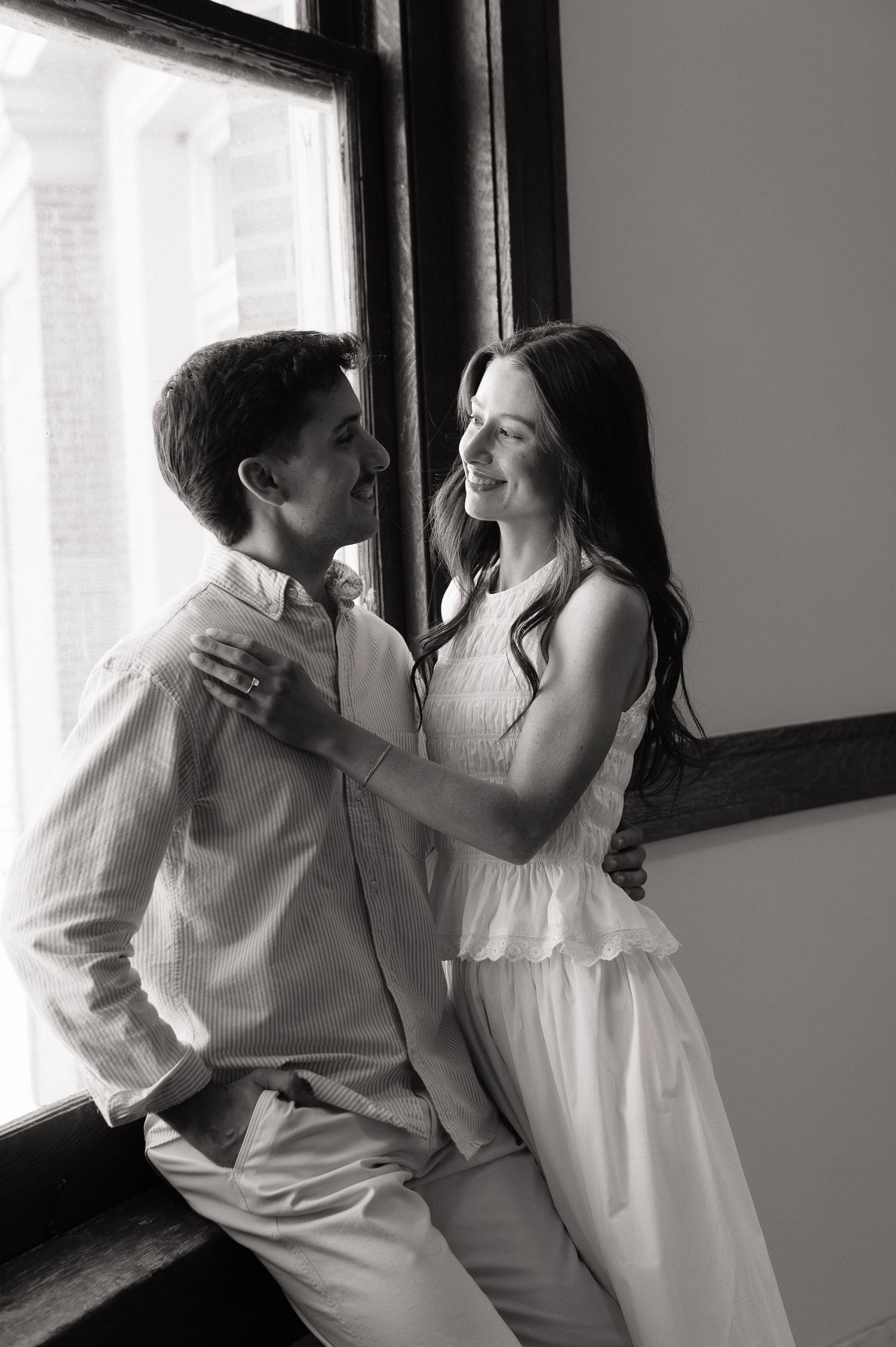 Black and white photo of a couple embracing and smiling by a window, woman in white dress, man in light shirt.