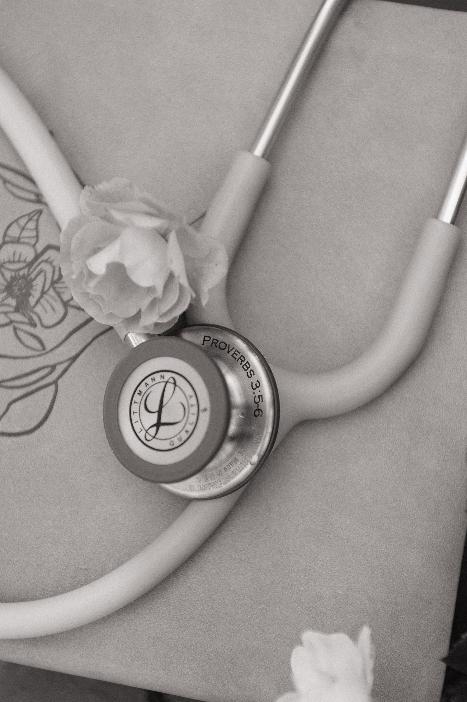 Black and white photo of a Littmann stethoscope with white flowers resting on a book inscribed with Proverbs 3:5-6.