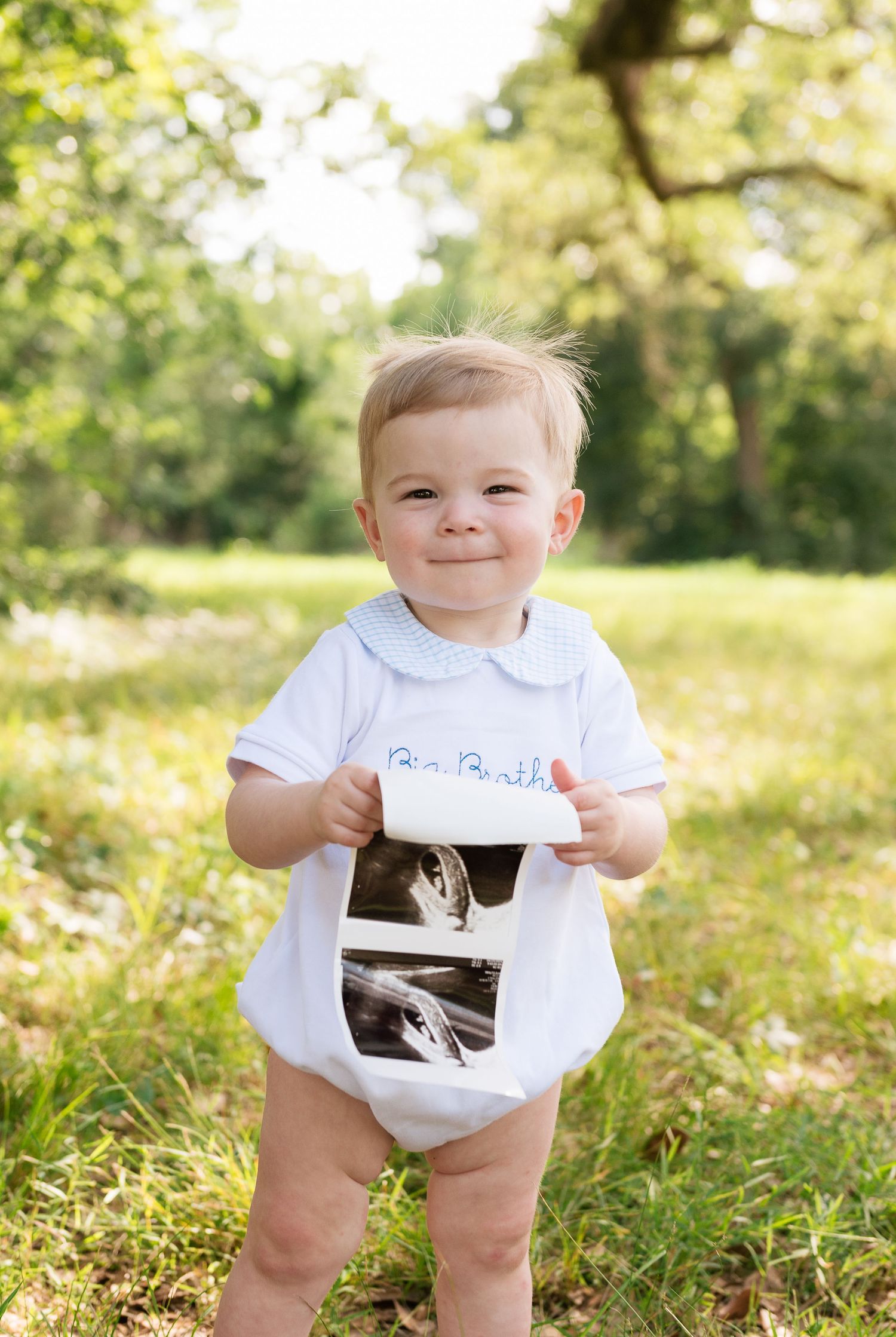 Smiling toddler in a white Big Brother onesie holds an ultrasound photo outdoors in a sunny green park.