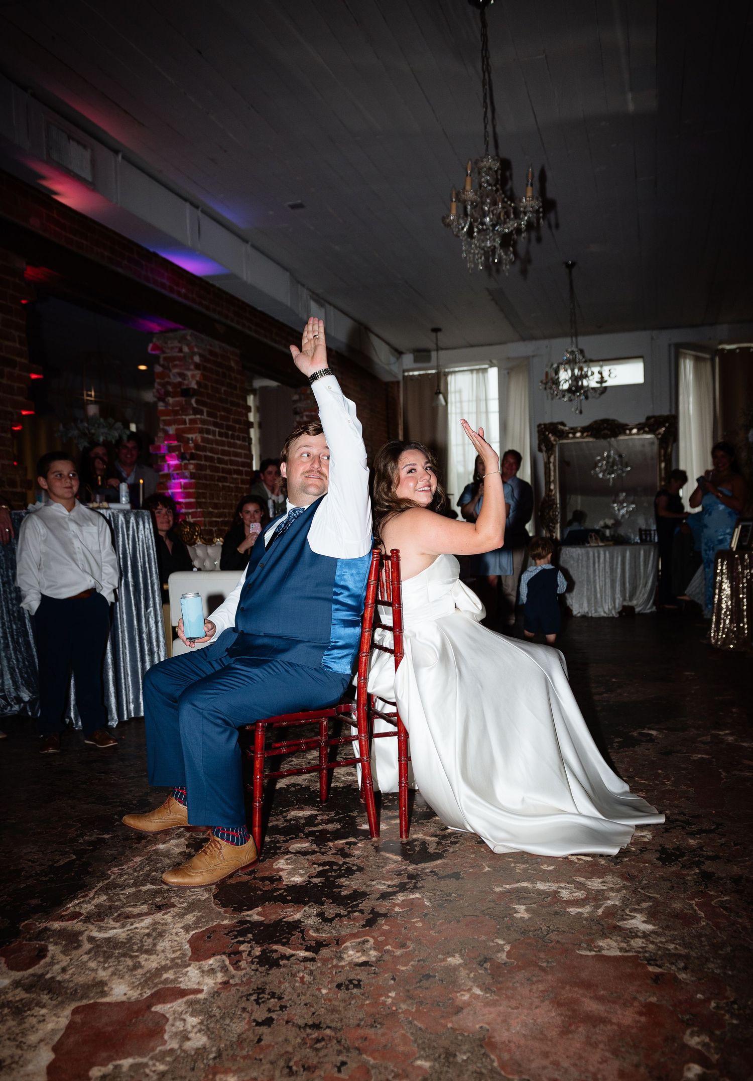 Bride and groom raise hands joyfully while seated on red chairs during their wedding reception in an elegant venue.