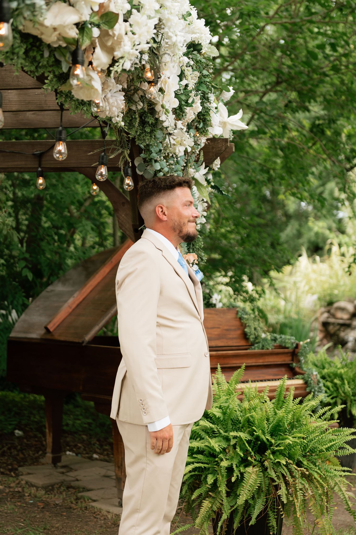 Groom in beige suit stands at outdoor altar adorned with white flowers and greenery near a grand piano.