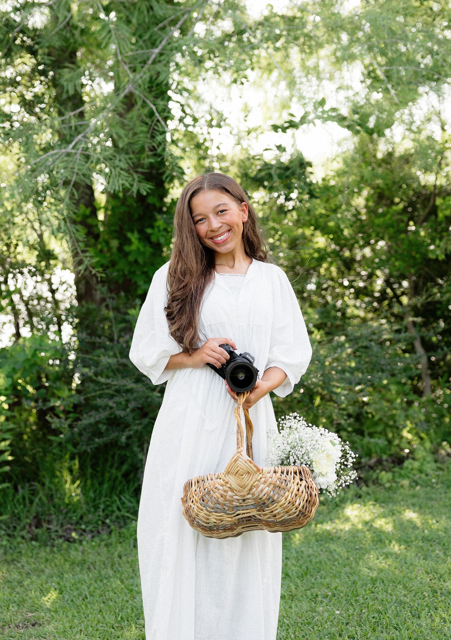 Woman in white dress holding camera and flower basket smiles outdoors among lush green trees.