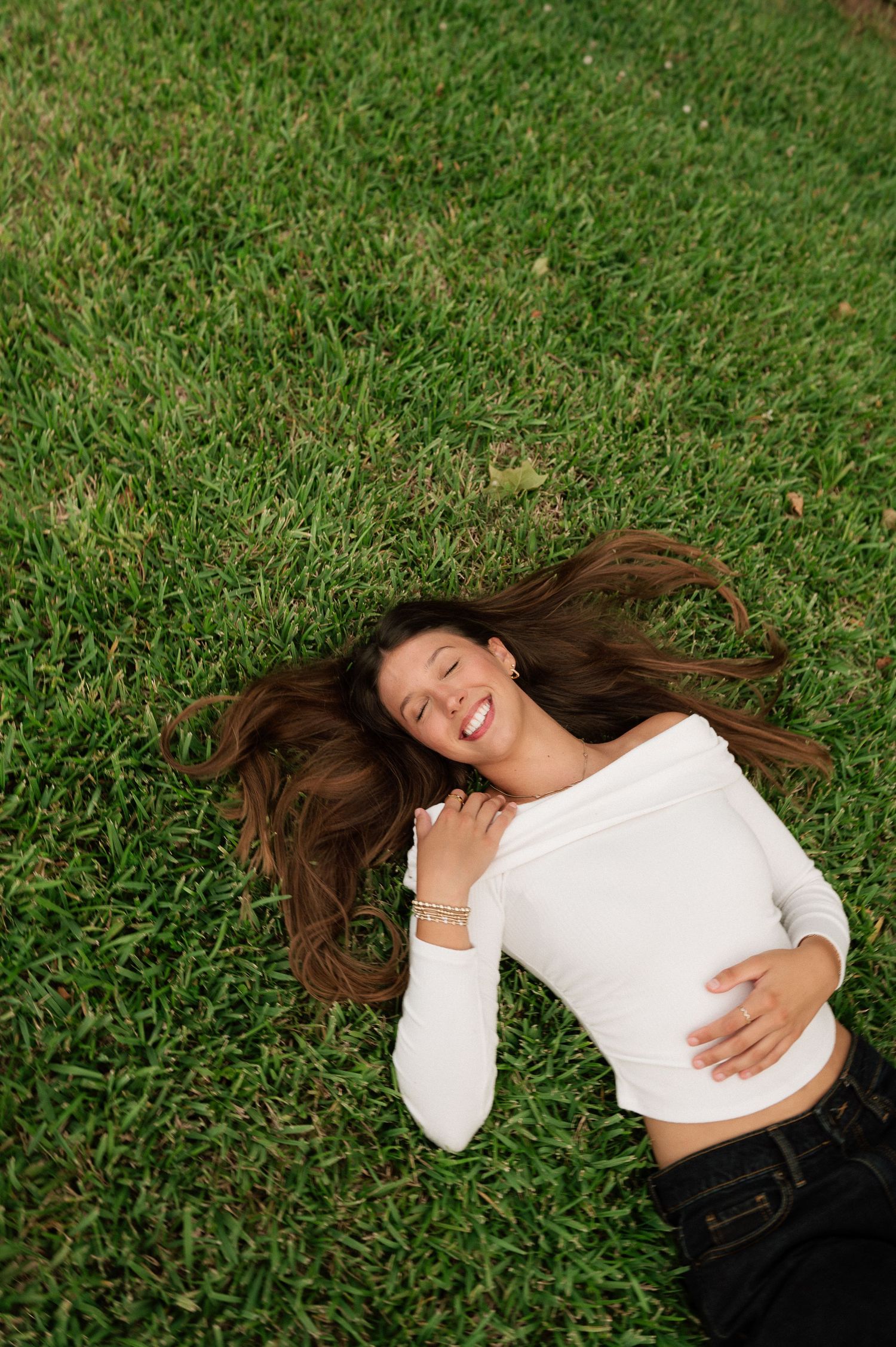 Smiling young woman with long brown hair lying on green grass, wearing a white off-shoulder top and dark jeans, eyes closed.