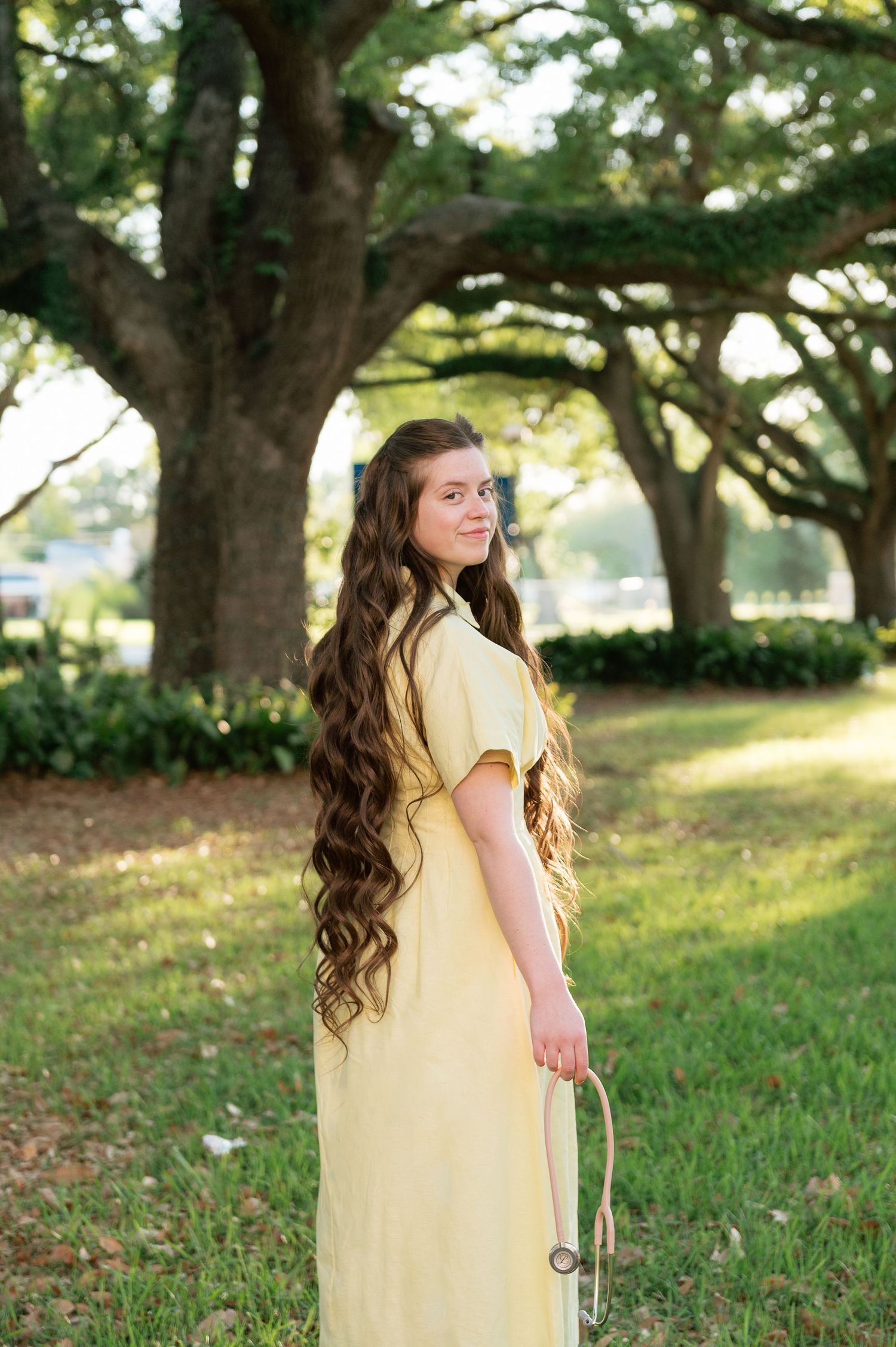 Young woman with long curly hair in a yellow dress stands outdoors among large oak trees on a green lawn.