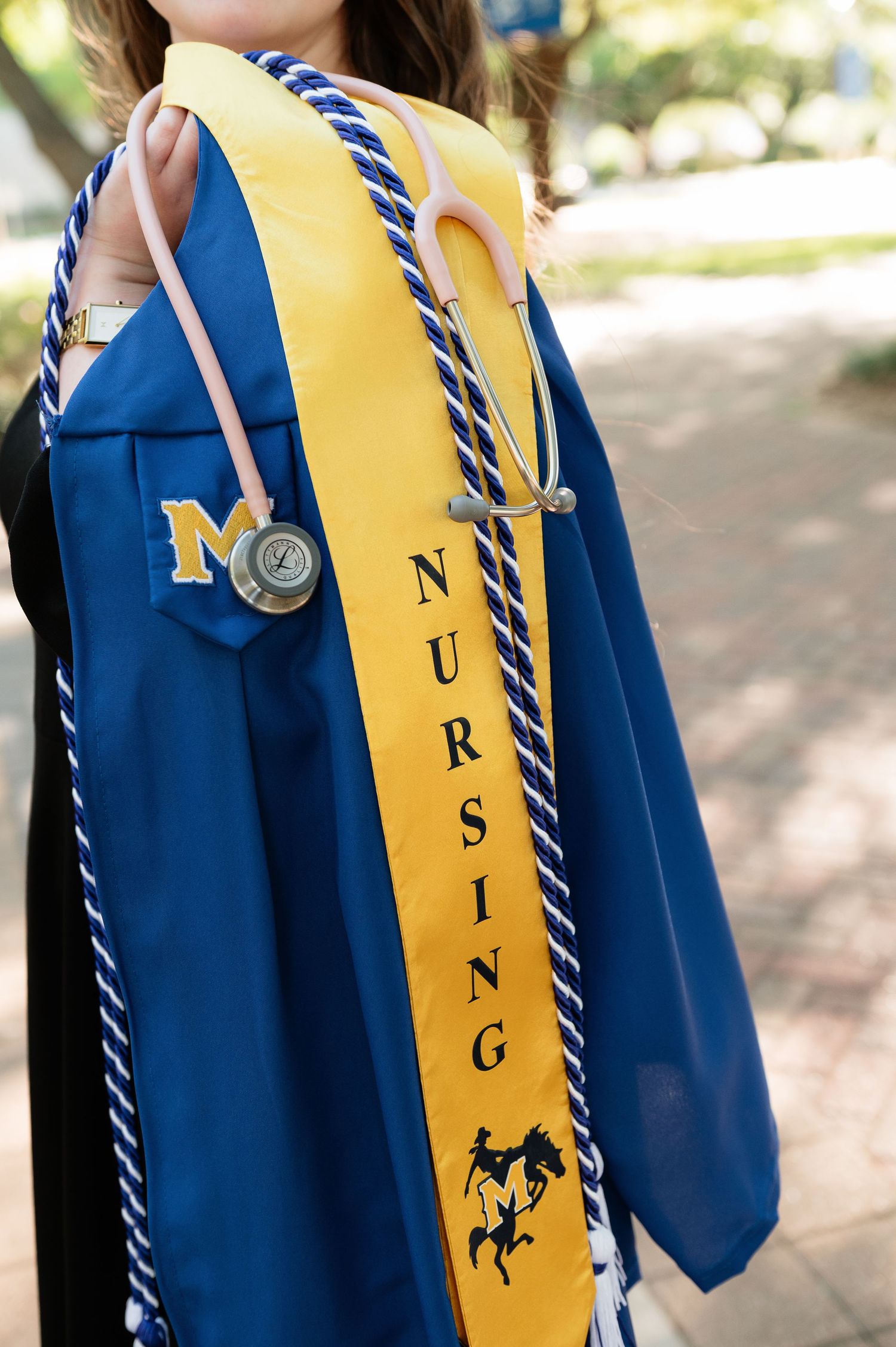 Nursing graduate in blue cap and gown with yellow sash, honor cords, and stethoscope at outdoor ceremony.