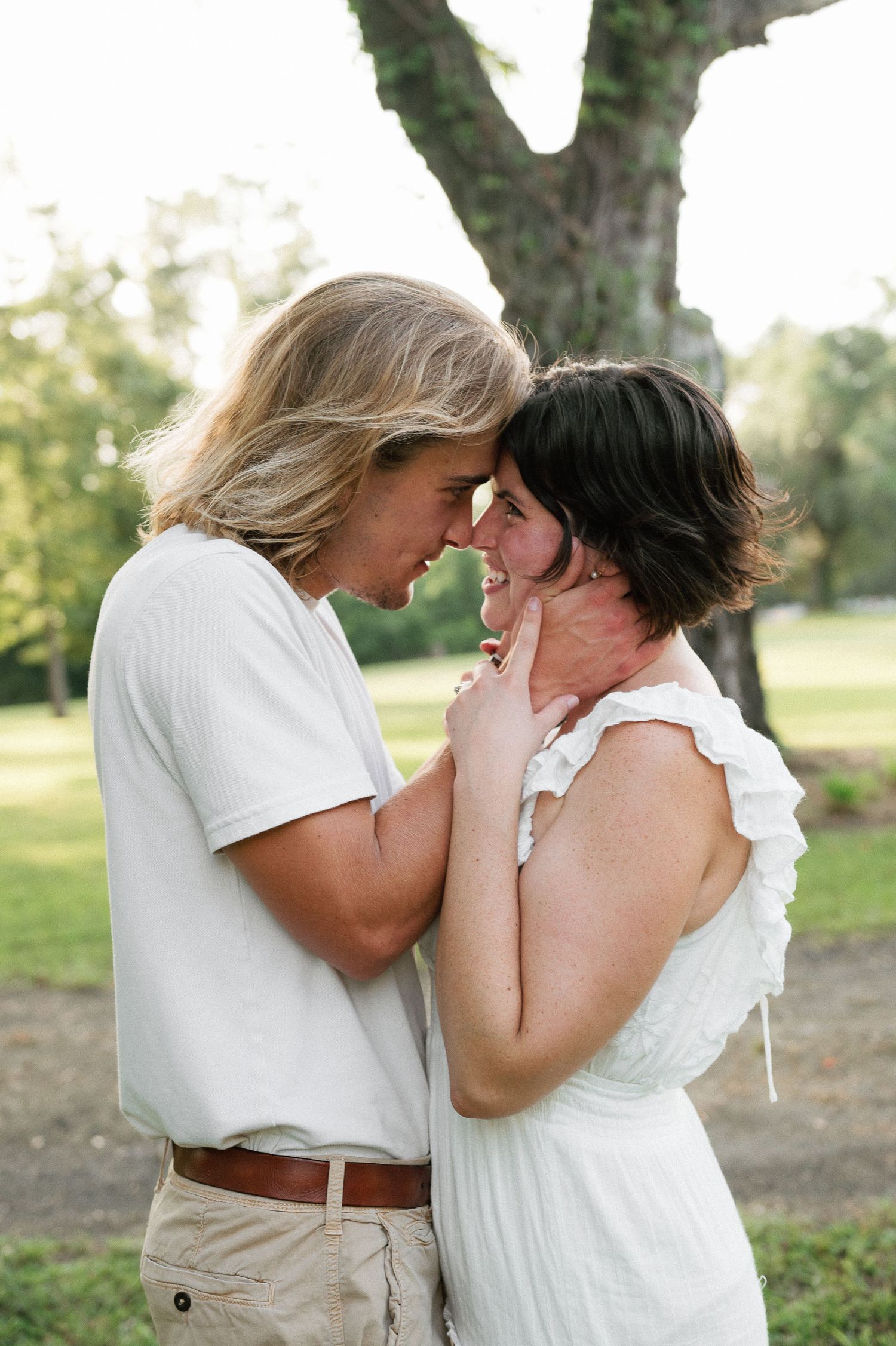 Romantic couple in white outfits touching foreheads tenderly outdoors near a large tree in soft natural light.