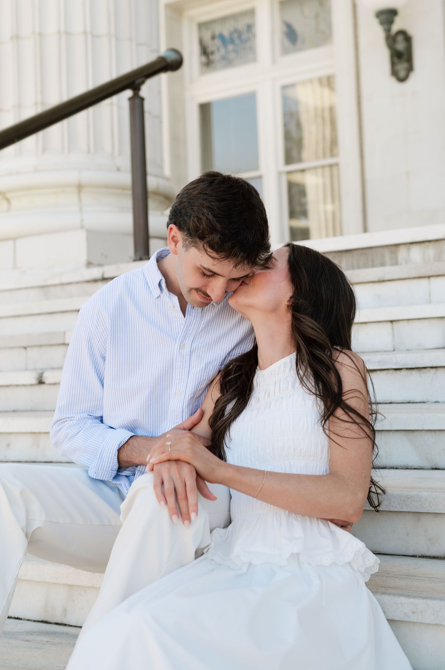 A couple shares a tender kiss on white marble steps, woman in white dress and man in light blue shirt.