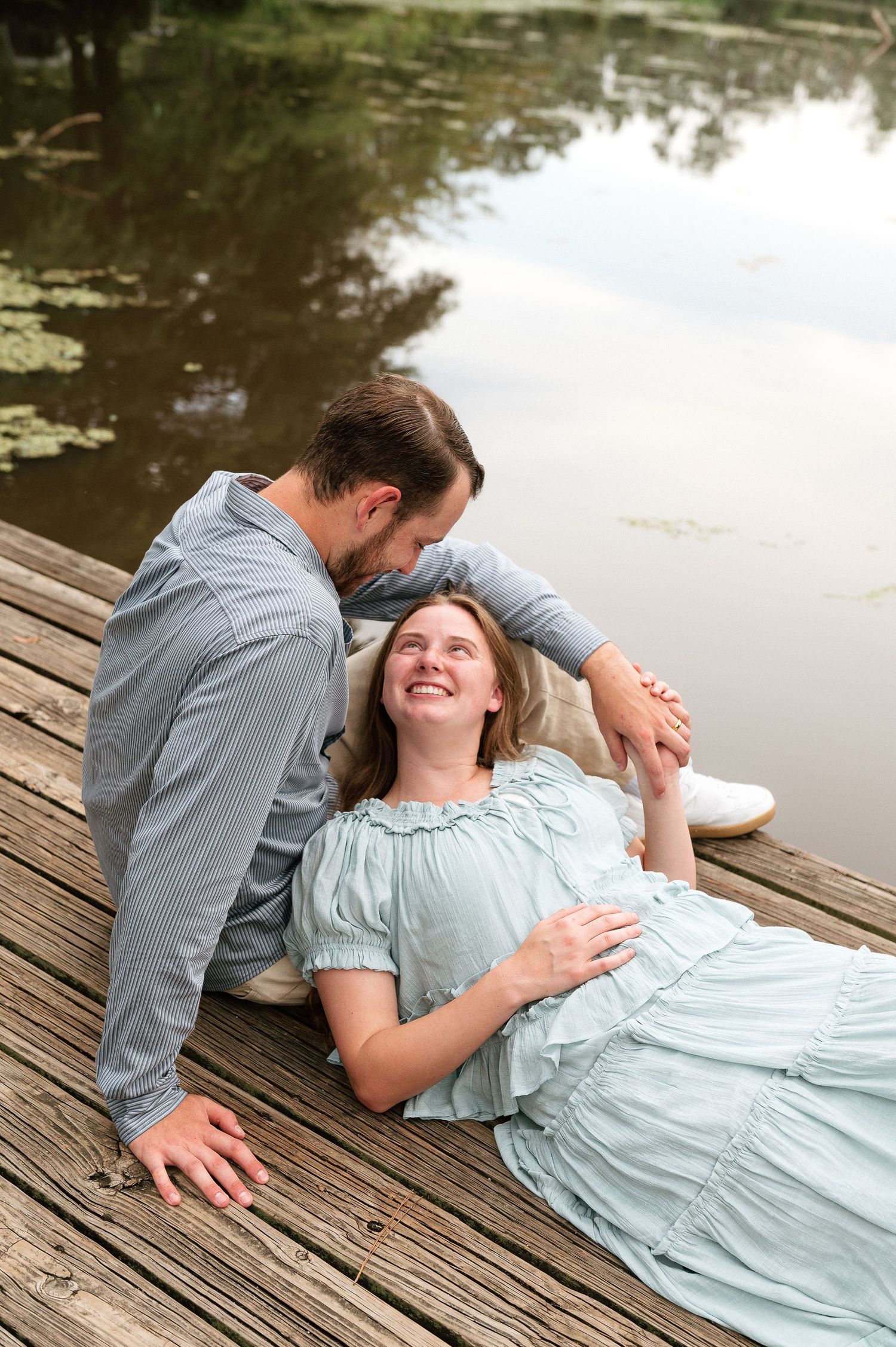 Pregnant woman in light blue dress lying on wooden dock, smiling up at man beside her near a peaceful pond.
