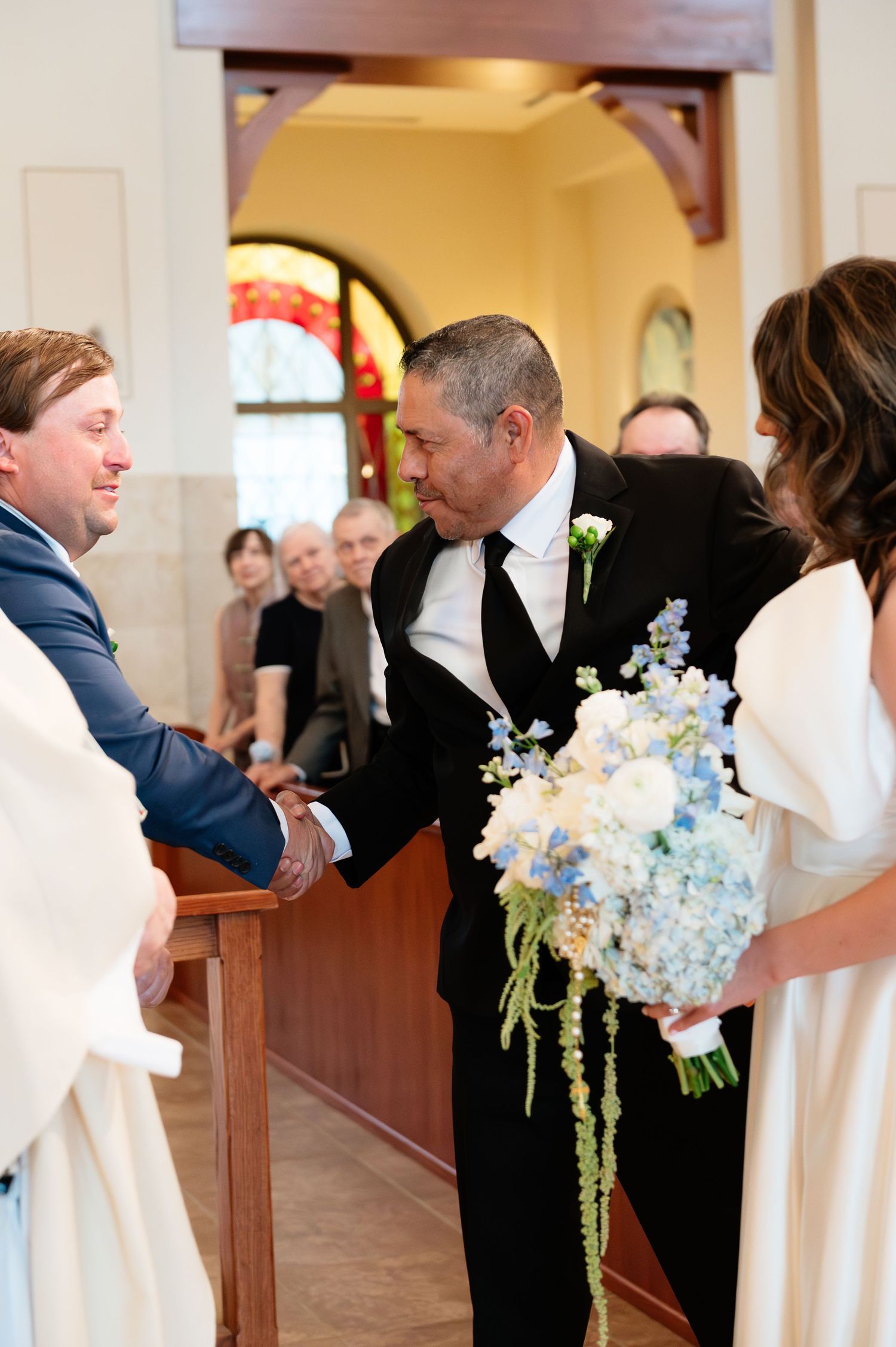 Groom shaking hands with guest while bride holds blue and white bouquet at church wedding.