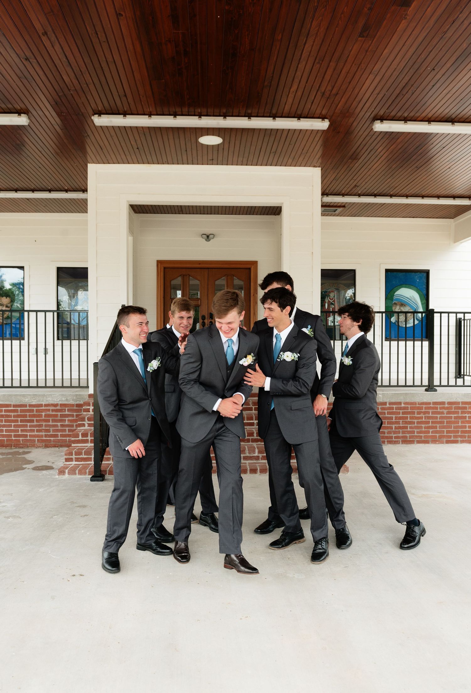 Groomsmen in gray suits with blue ties gather around the groom outside a brick building, laughing together.